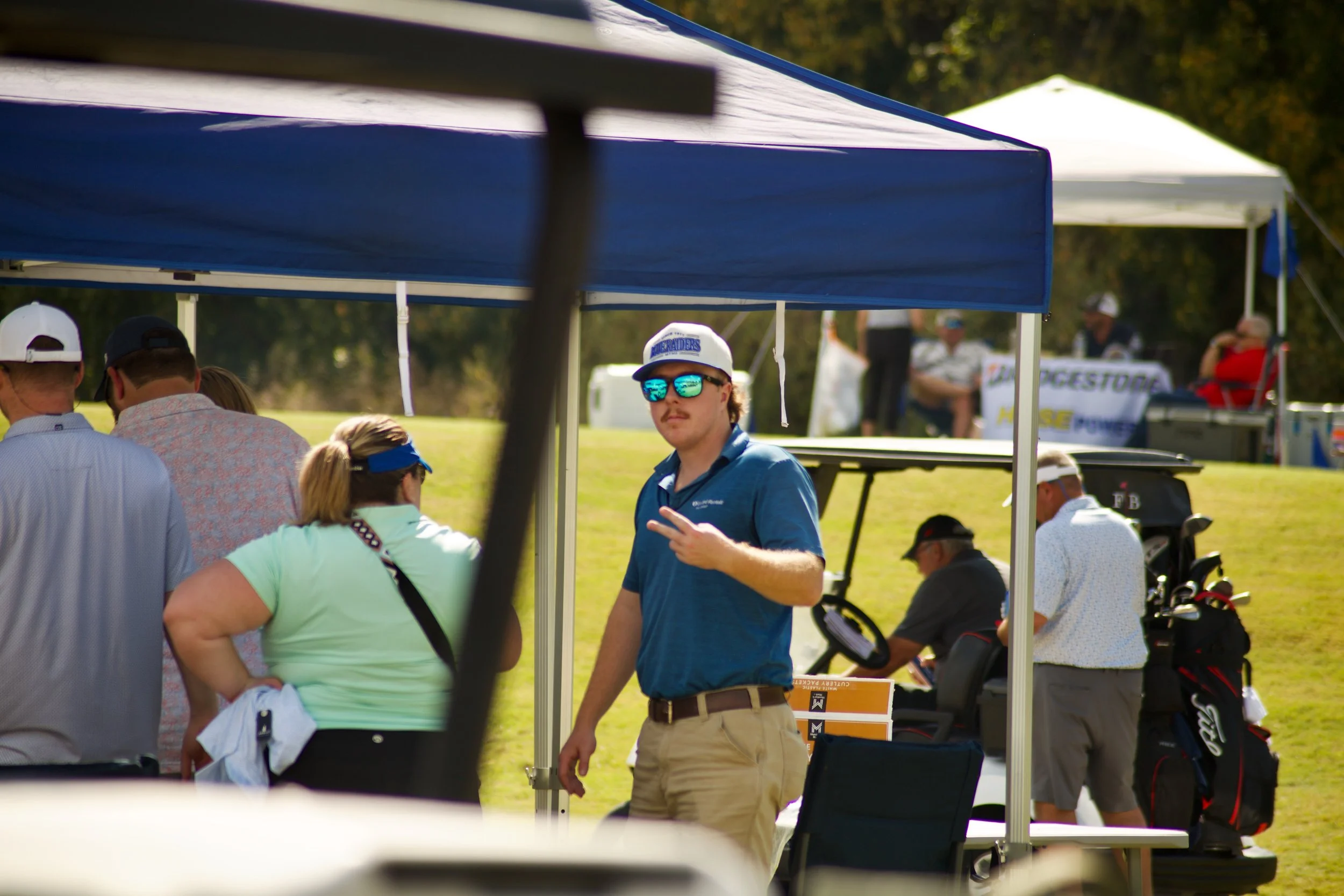 People gather outdoors at a golf event with tents, golf bags, and a man in the center wearing sunglasses and a blue shirt making a peace sign.