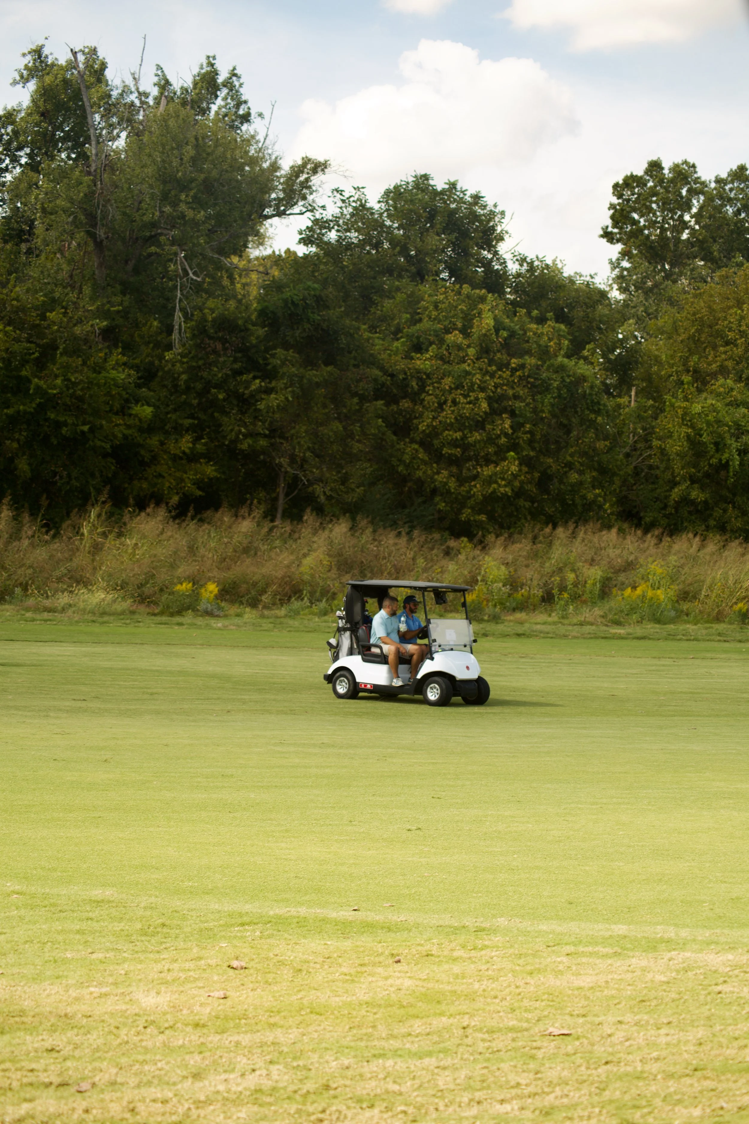 Two men in a golf cart on a golf course, with dense trees in the background and blue sky with clouds.
