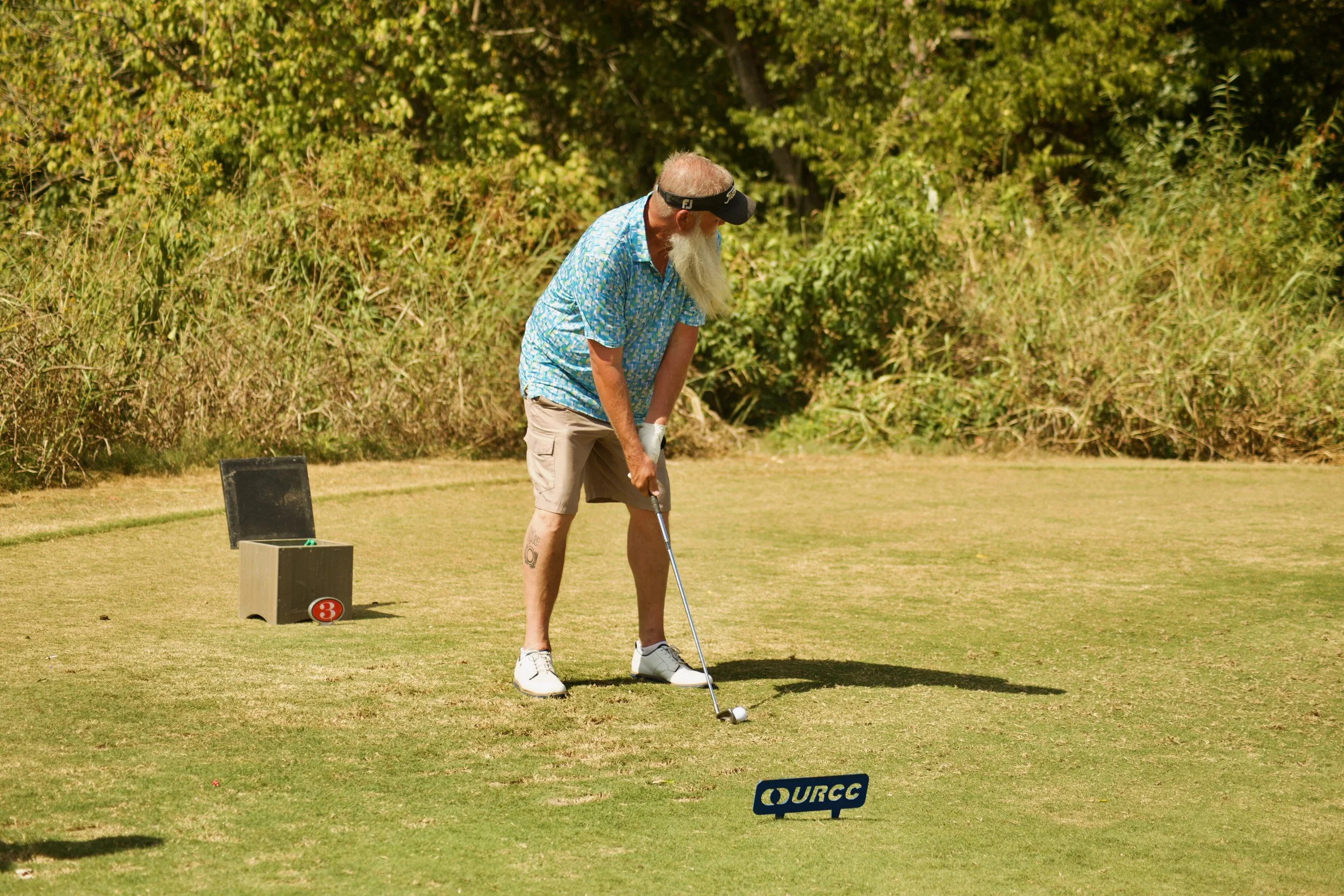 An older man with a long white beard, wearing a colorful short-sleeve shirt, light shorts, sneakers, a black visor, and gloves, is preparing to hit a golf ball on a grassy course surrounded by green trees.