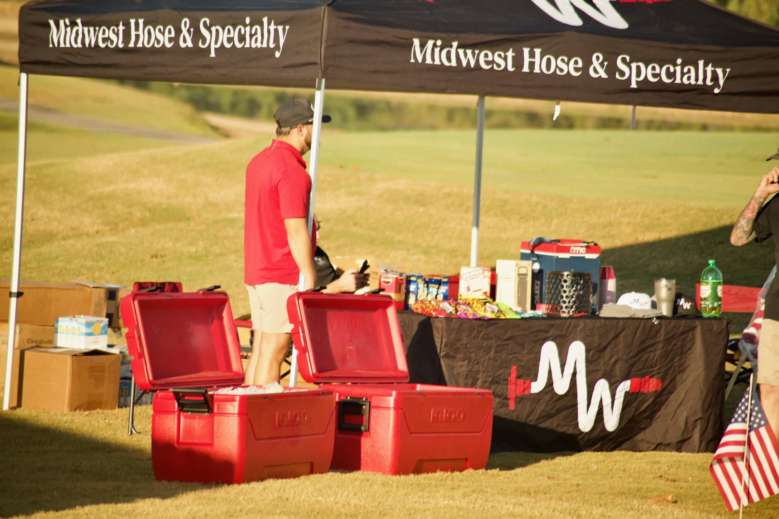 Outdoor booth with a black canopy advertising 'Midwest Hose & Specialty.' Man in red shirt and tan shorts looking at items on the table. Red coolers at the front, various boxes and items on the table, and grass field in background.