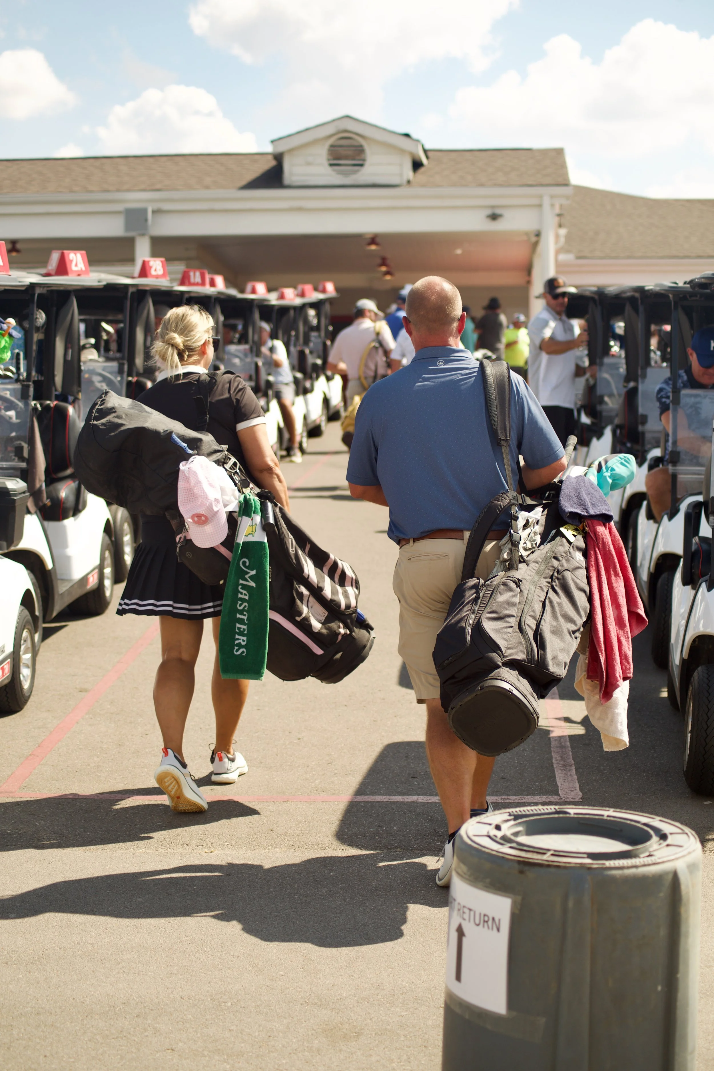 People walking with golf clubs and bags near golf carts at a golf course entrance on a sunny day