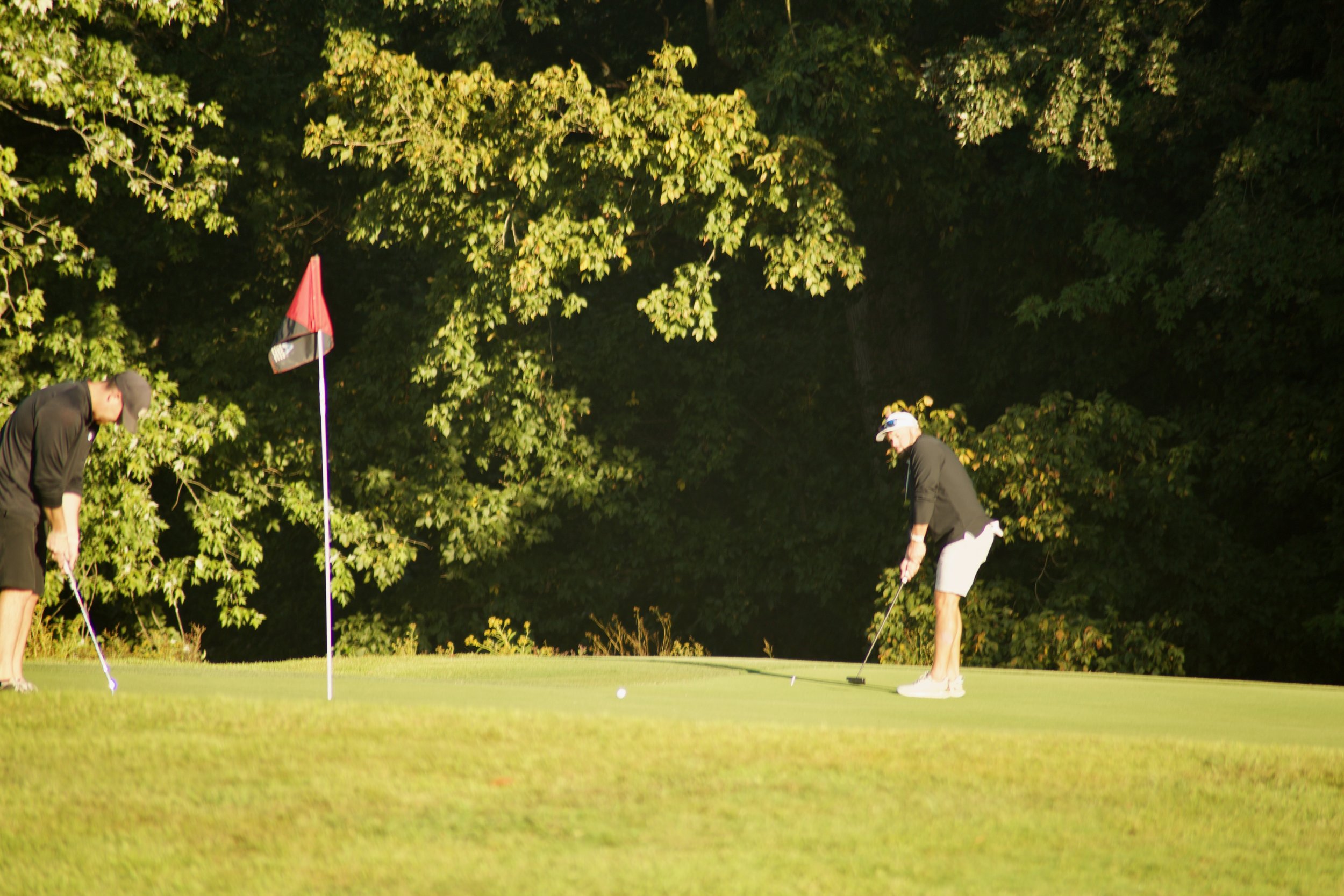 Two women and a man playing golf on a green course with trees in the background.