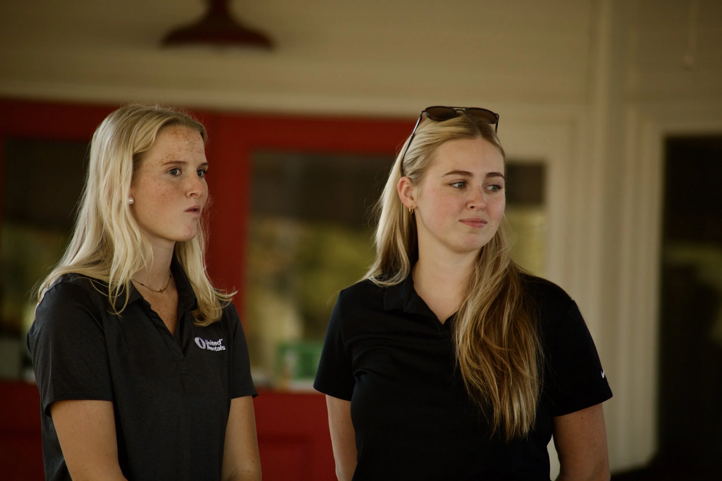 Two young women wearing black uniforms with United Rentals logos stand indoors with neutral expressions.