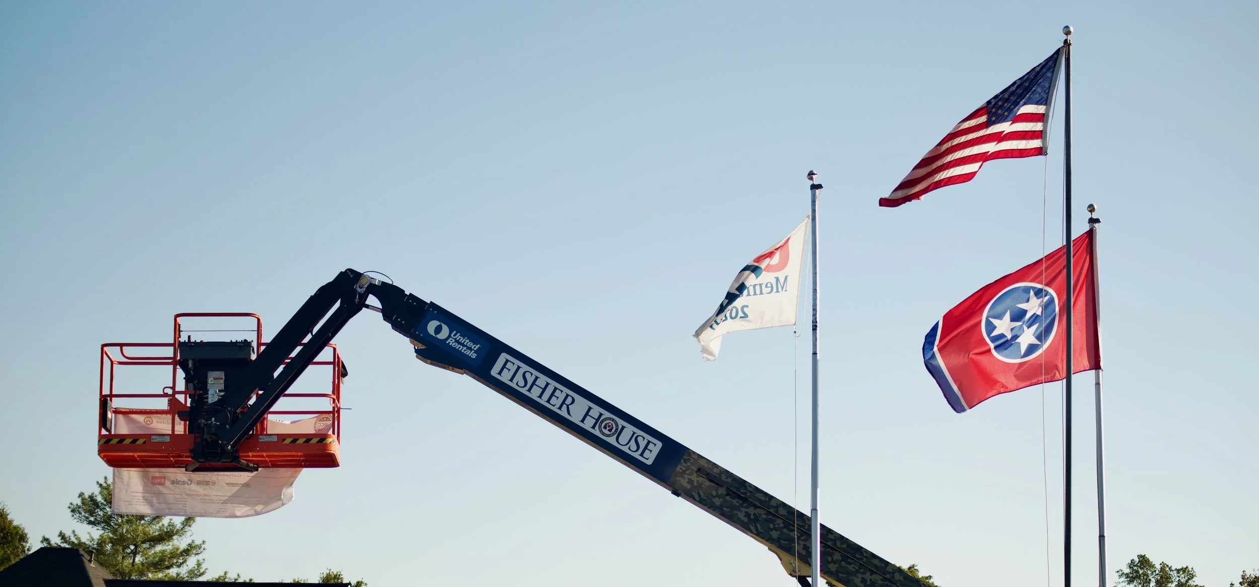 A crane labeled 'Fisher House' lifting a large banner with text, and three flags (U.S. flag, Tennessee flag, and a white flag) flying against a clear blue sky.