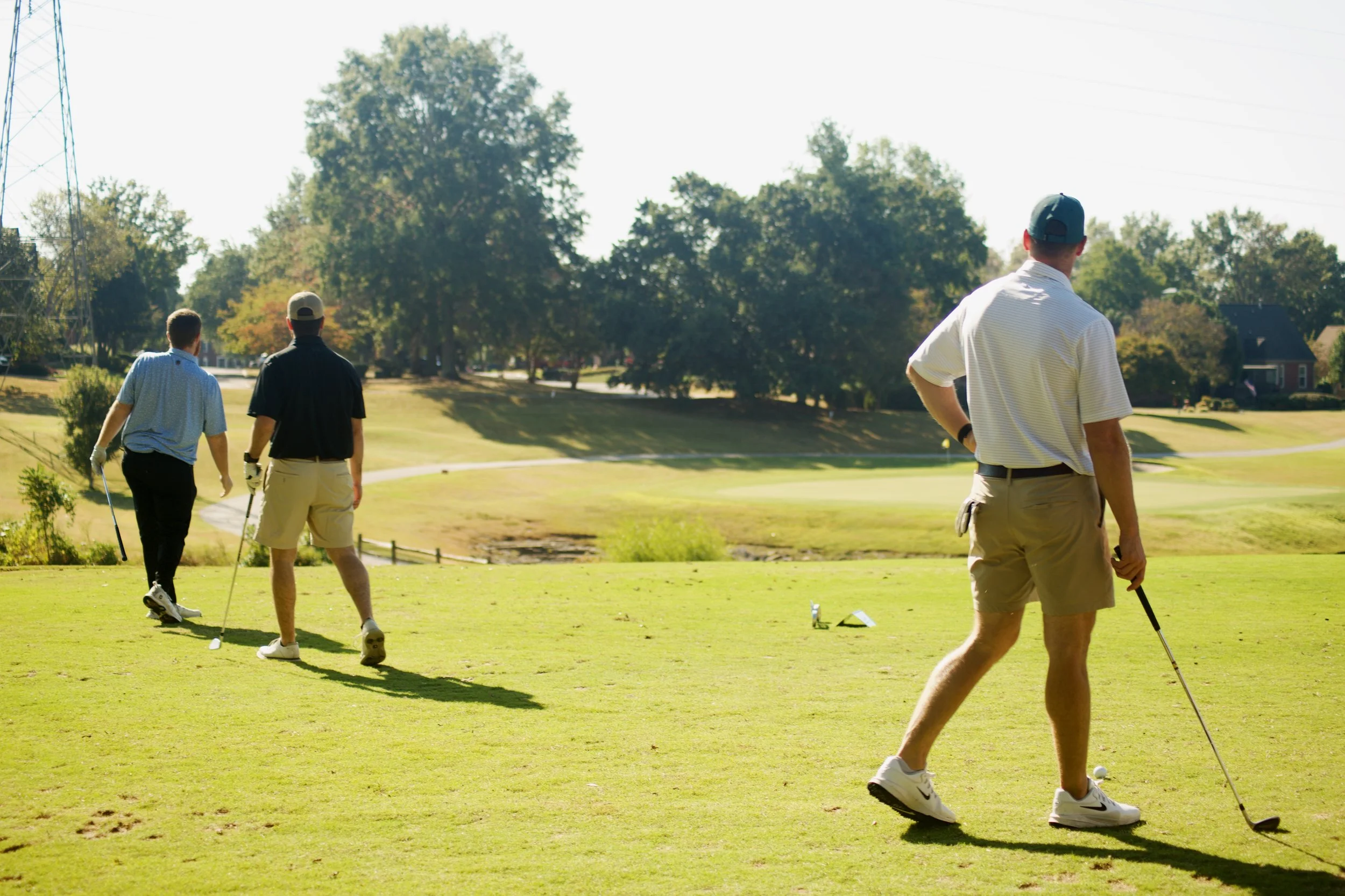 Three men on a golf course with trees and houses in the background, walking and preparing to hit golf balls. All are dressed in casual golf attire.