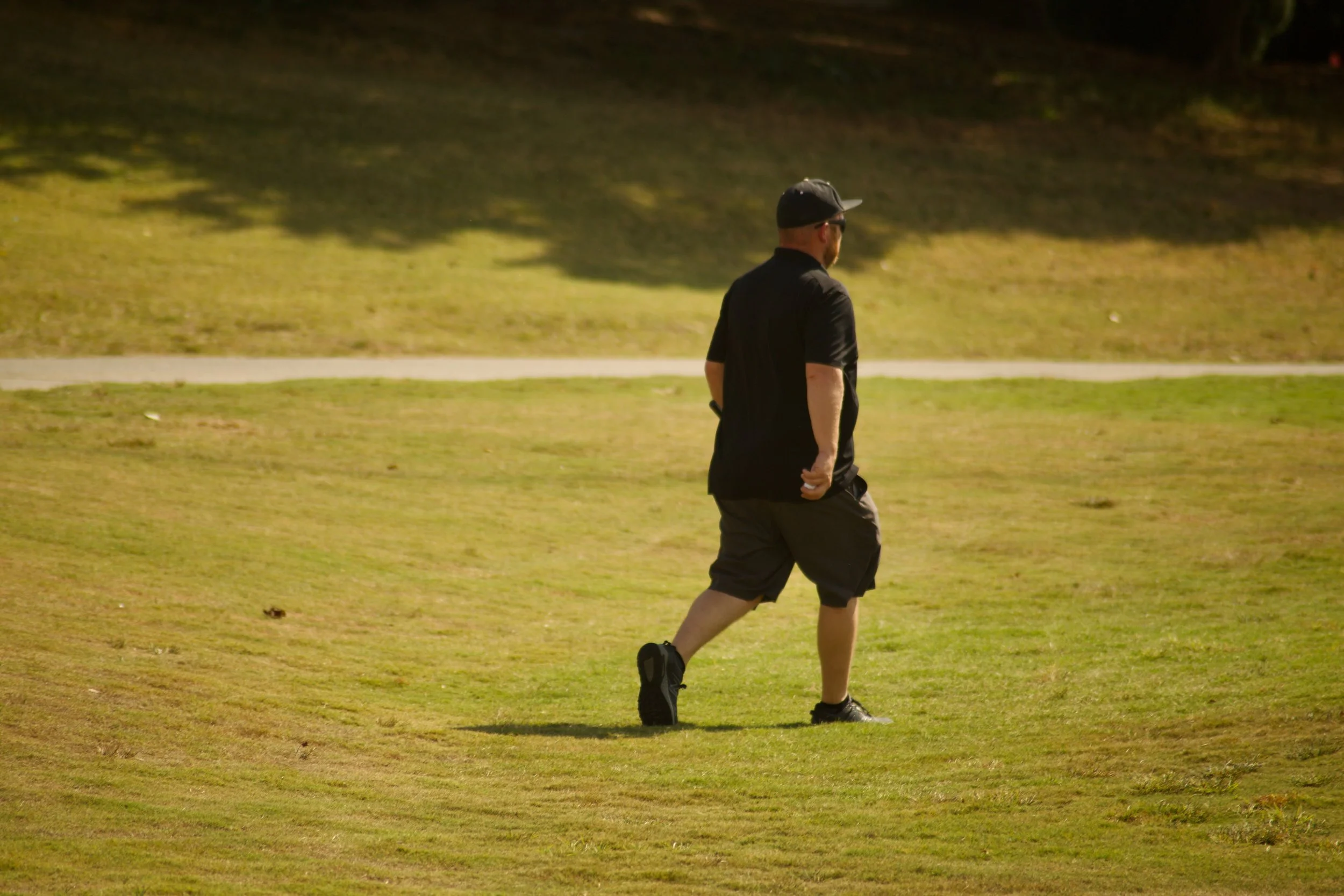 A man dressed in black, wearing a cap and sunglasses, walking on a grassy area in a park.