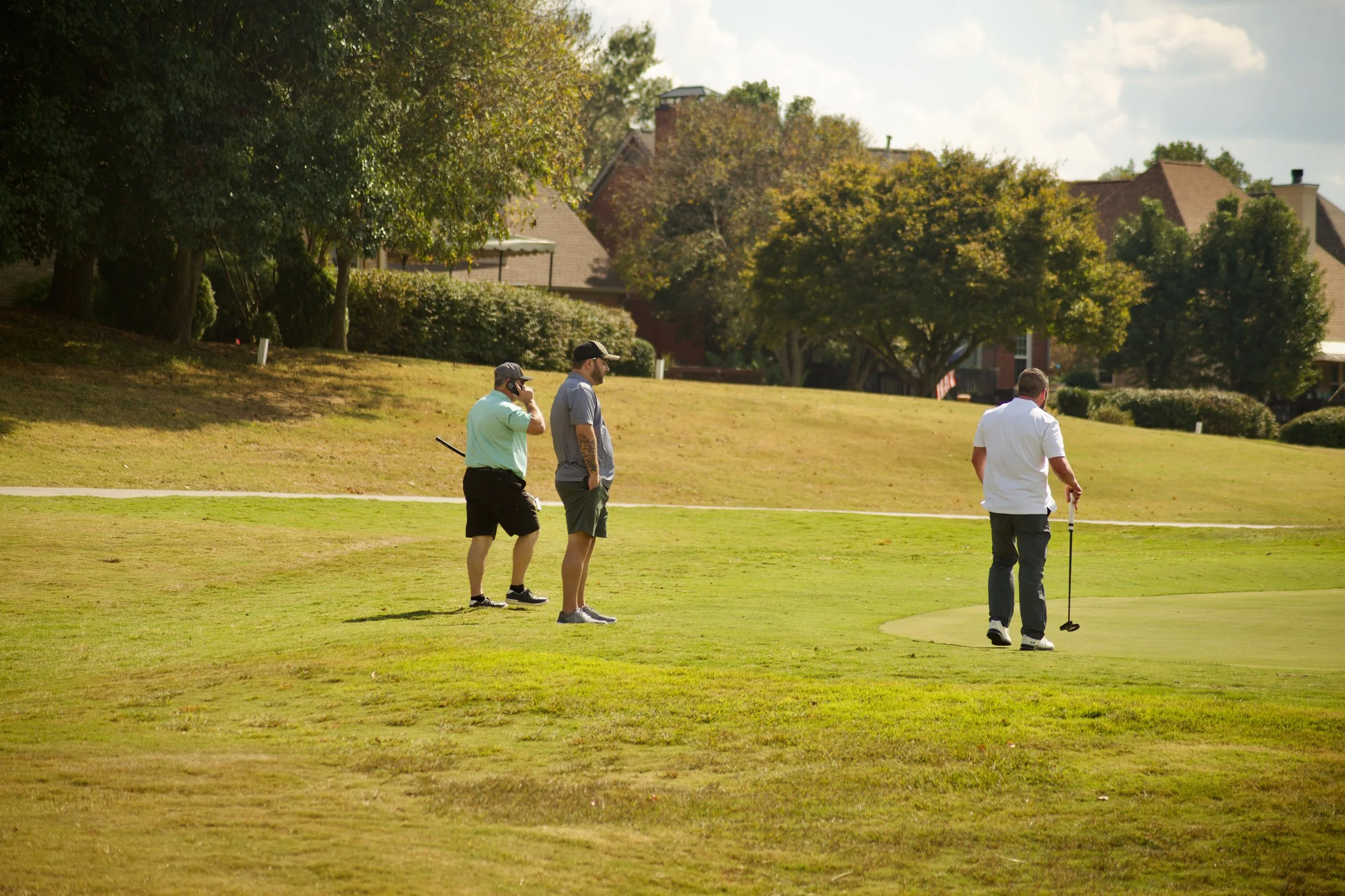 Three men on a golf course; one preparing to putt, and the others standing nearby watching.