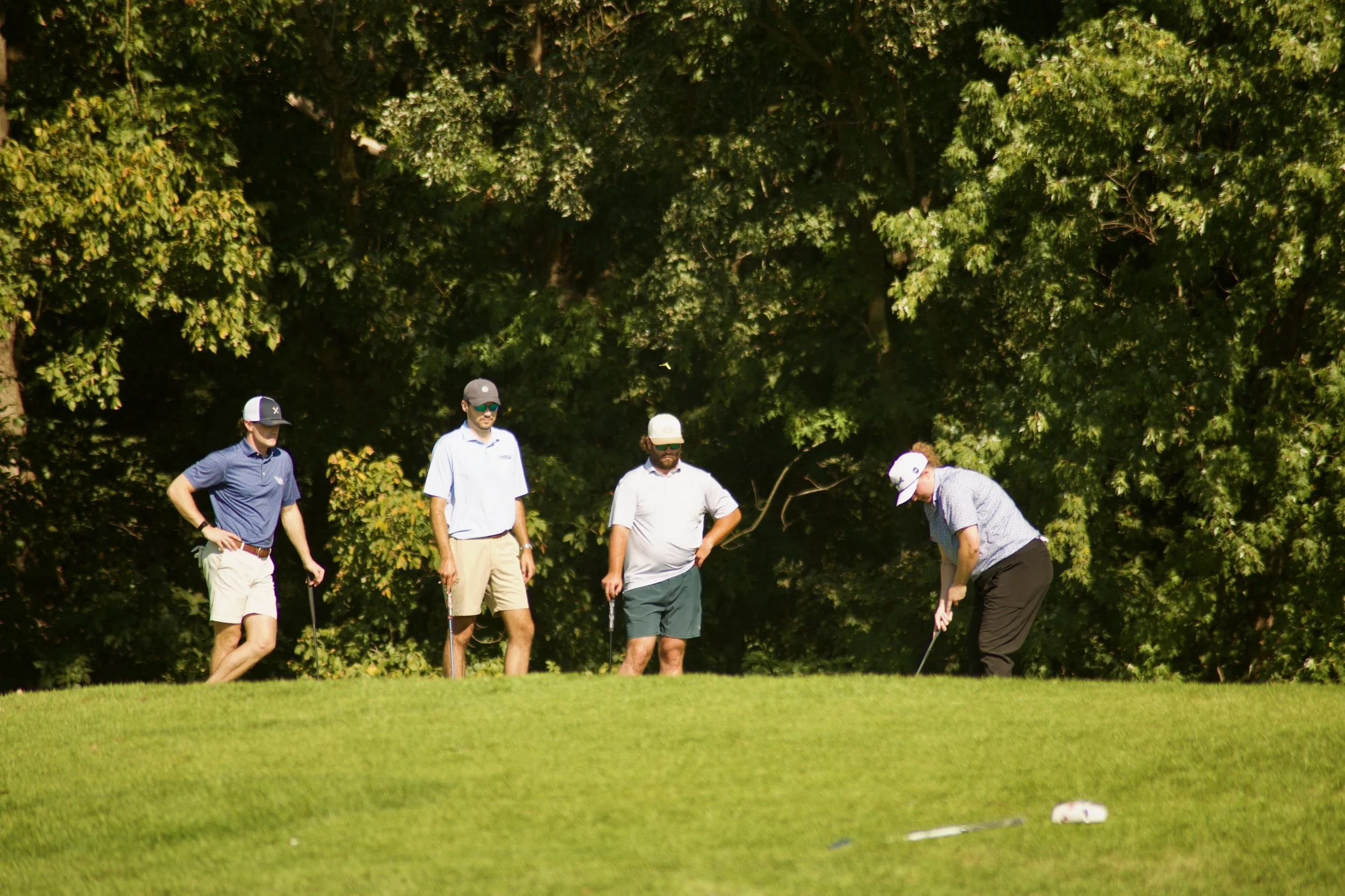 Four men playing golf on a lush green course, with tall trees in the background, some of the men are preparing to hit the ball.