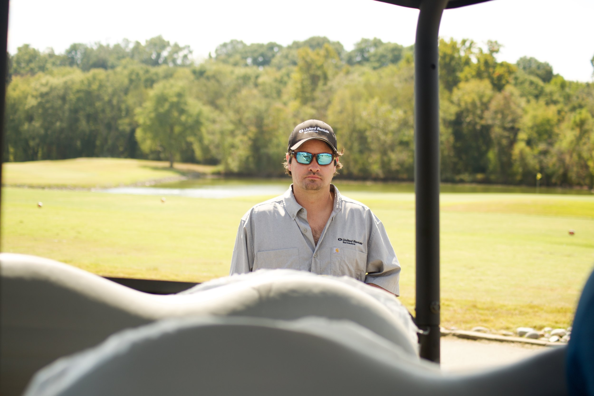 Person standing outdoors on a grassy area, wearing sunglasses, a cap, and a work shirt, with trees and a pond in the background, seen from inside a golf cart.