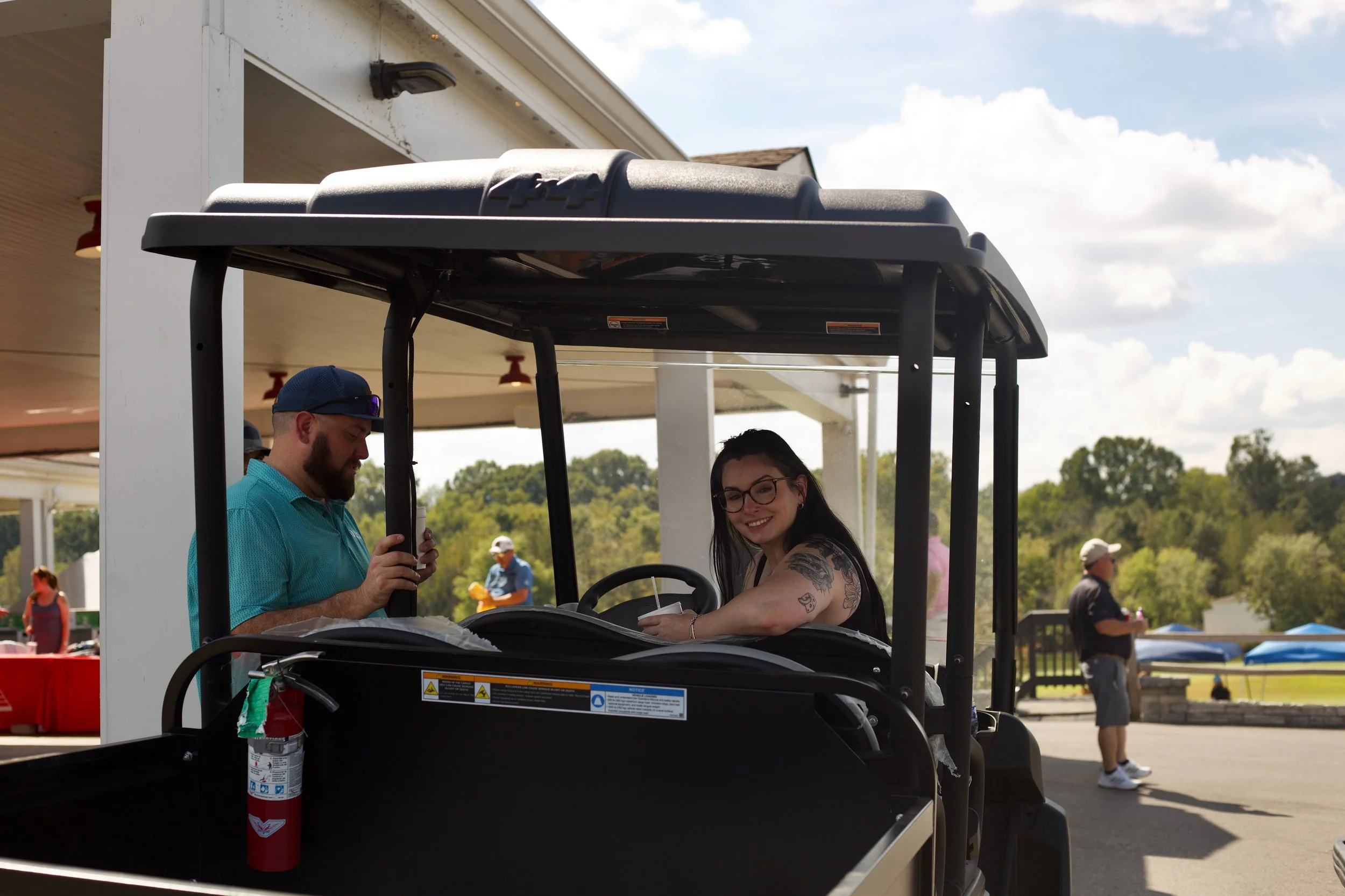 A woman with tattoos on her arm smiling while sitting in a golf cart, with a man standing beside her wearing a hat and sunglasses, holding a drink. The scene appears to be outdoors at a park or event with other people walking around in the background