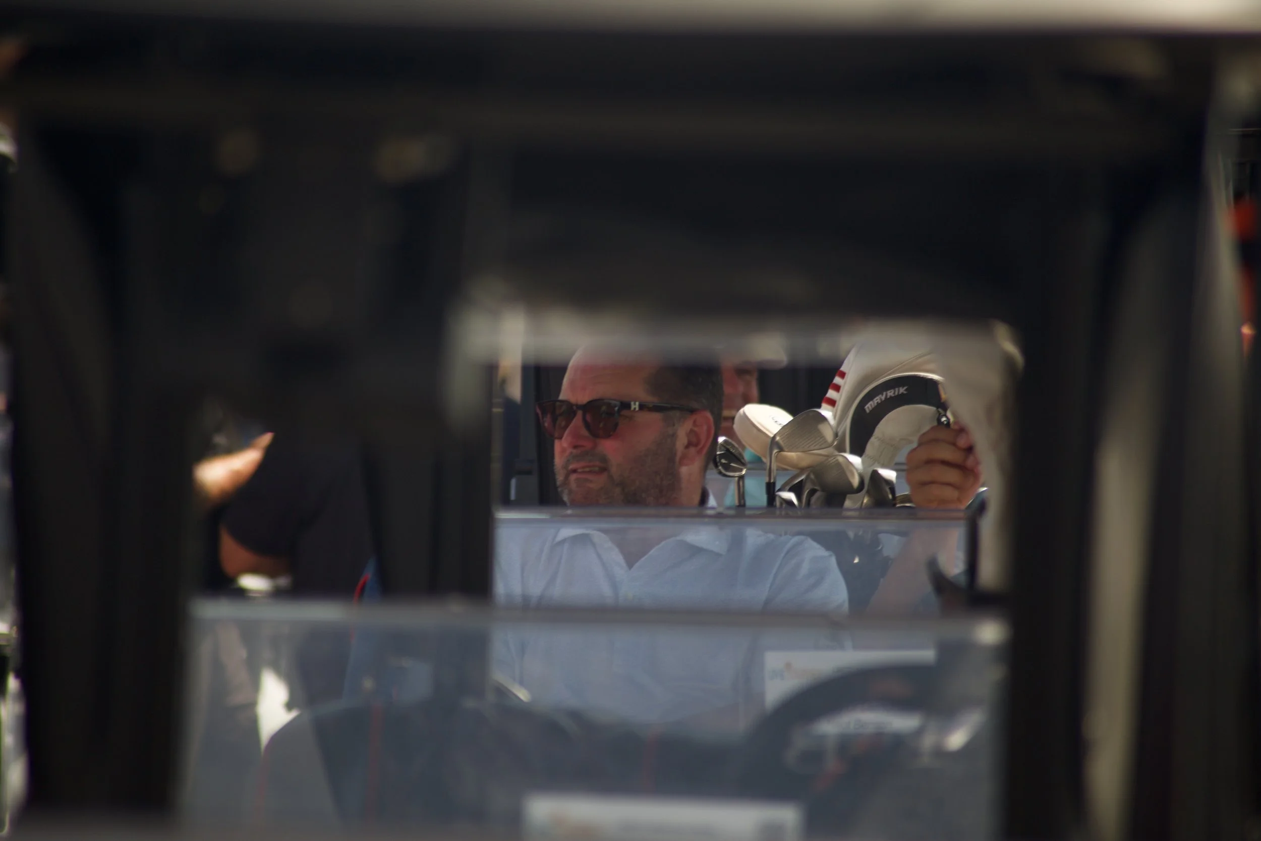 A man with a beard and glasses looking at golf clubs inside a vehicle, possibly a golf cart, through a rear window.