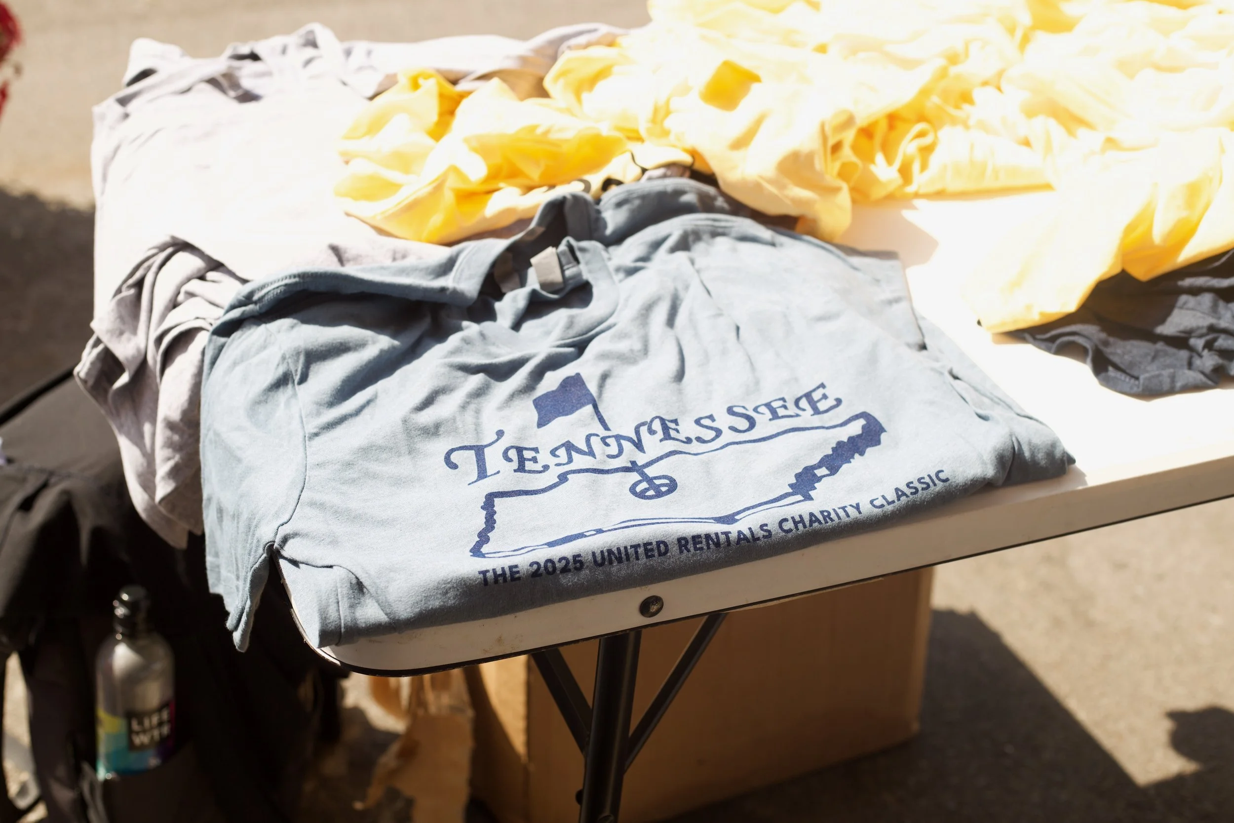 Clothes, including a gray T-shirt with Tennessee theme and yellow fabric, are laid out on a table, with a gas cylinder partially visible underneath, indicating outdoor setting.