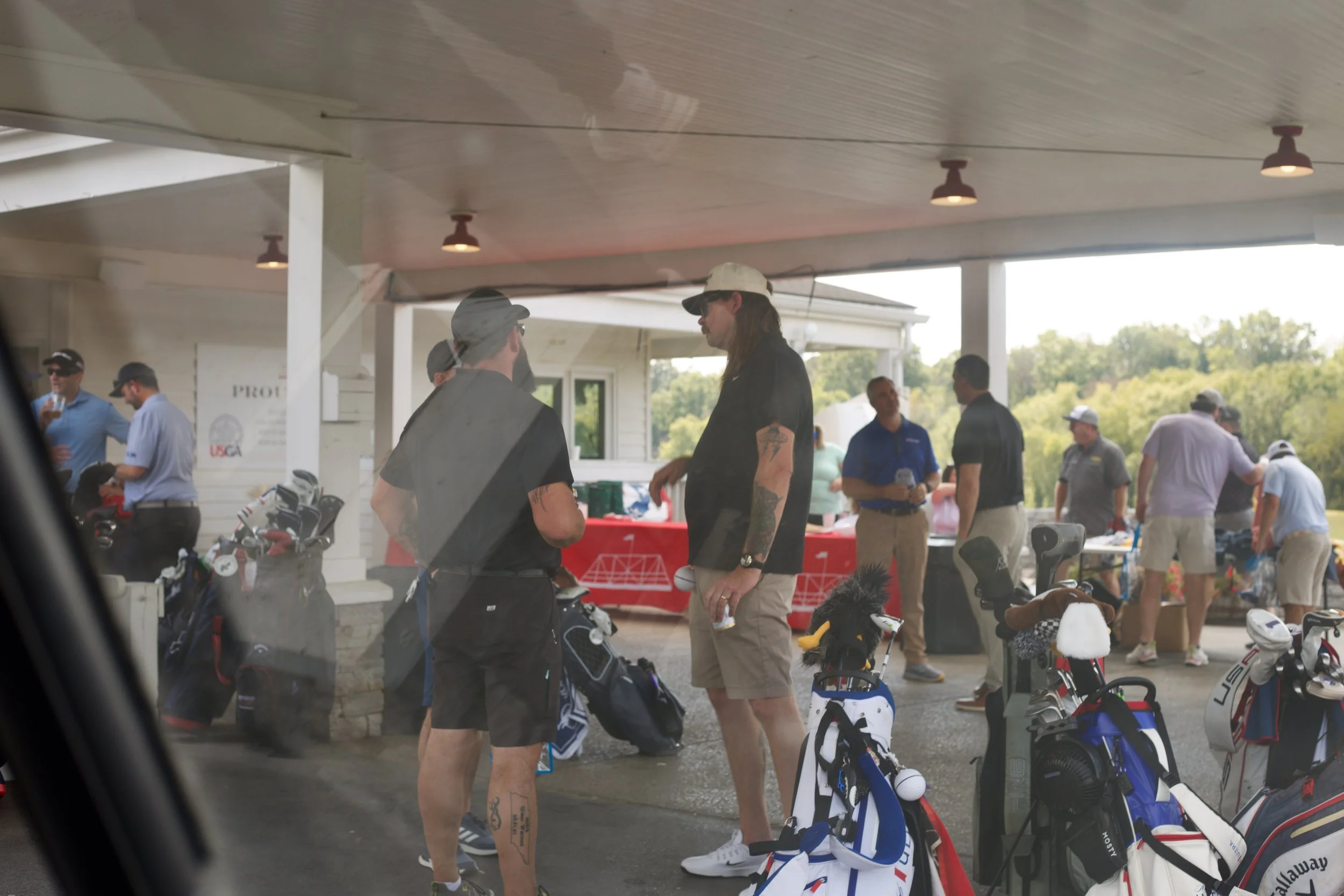 People at a golf practice area with golf bags and clubs, socializing and preparing for golf, outdoors under a covered area with trees in the background.