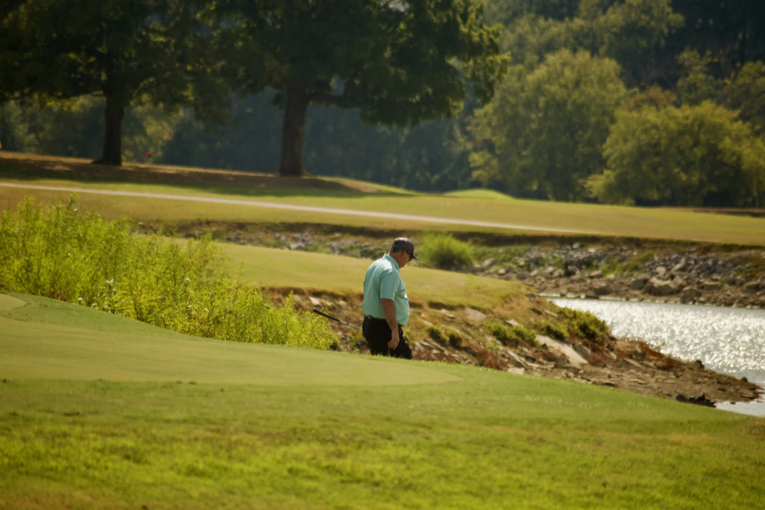 A man walking along a water's edge on a golf course, surrounded by lush green trees and grass, with a scenic landscape in the background.