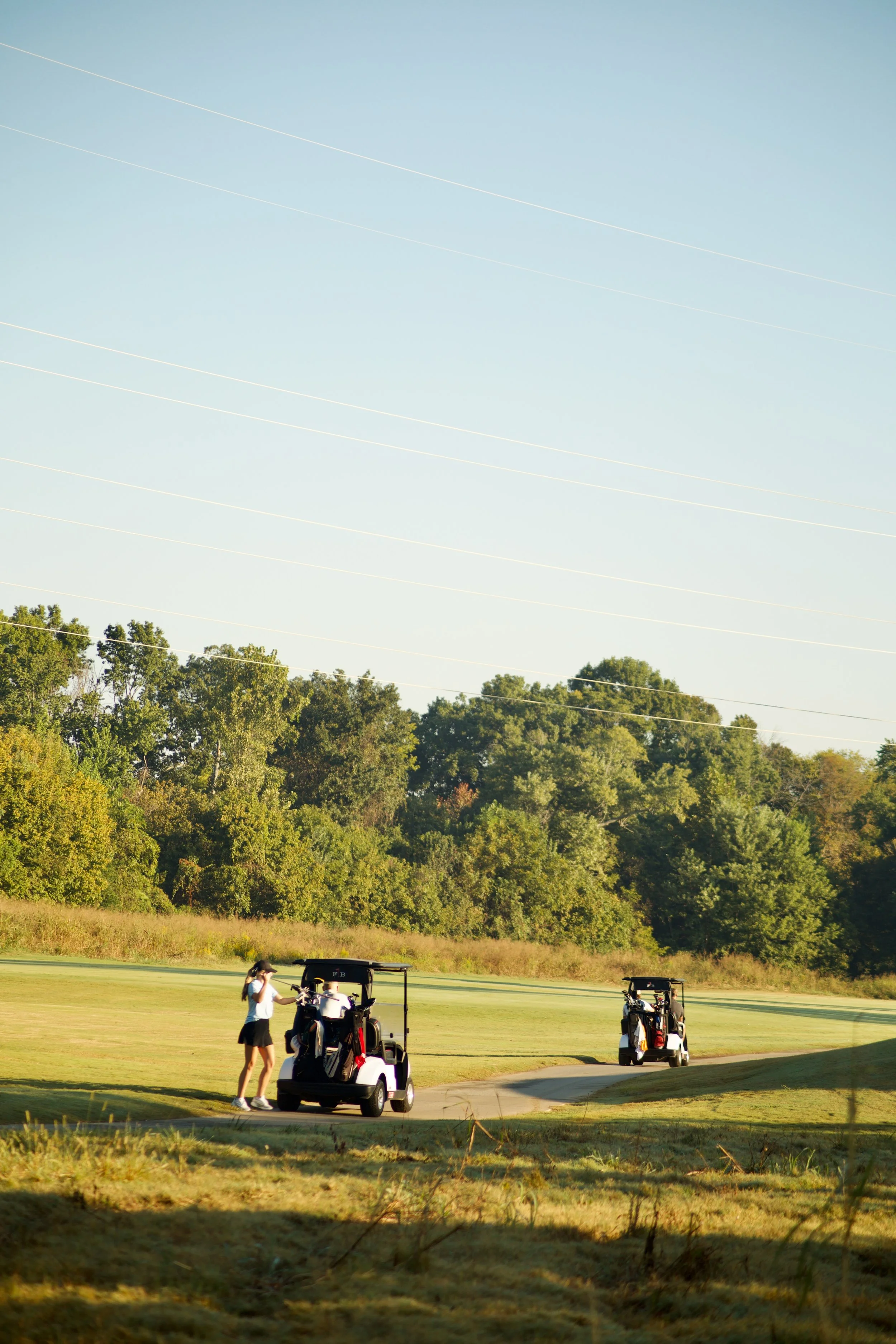 Two women and two golf carts on a golf course with trees and a clear blue sky, and power lines overhead.