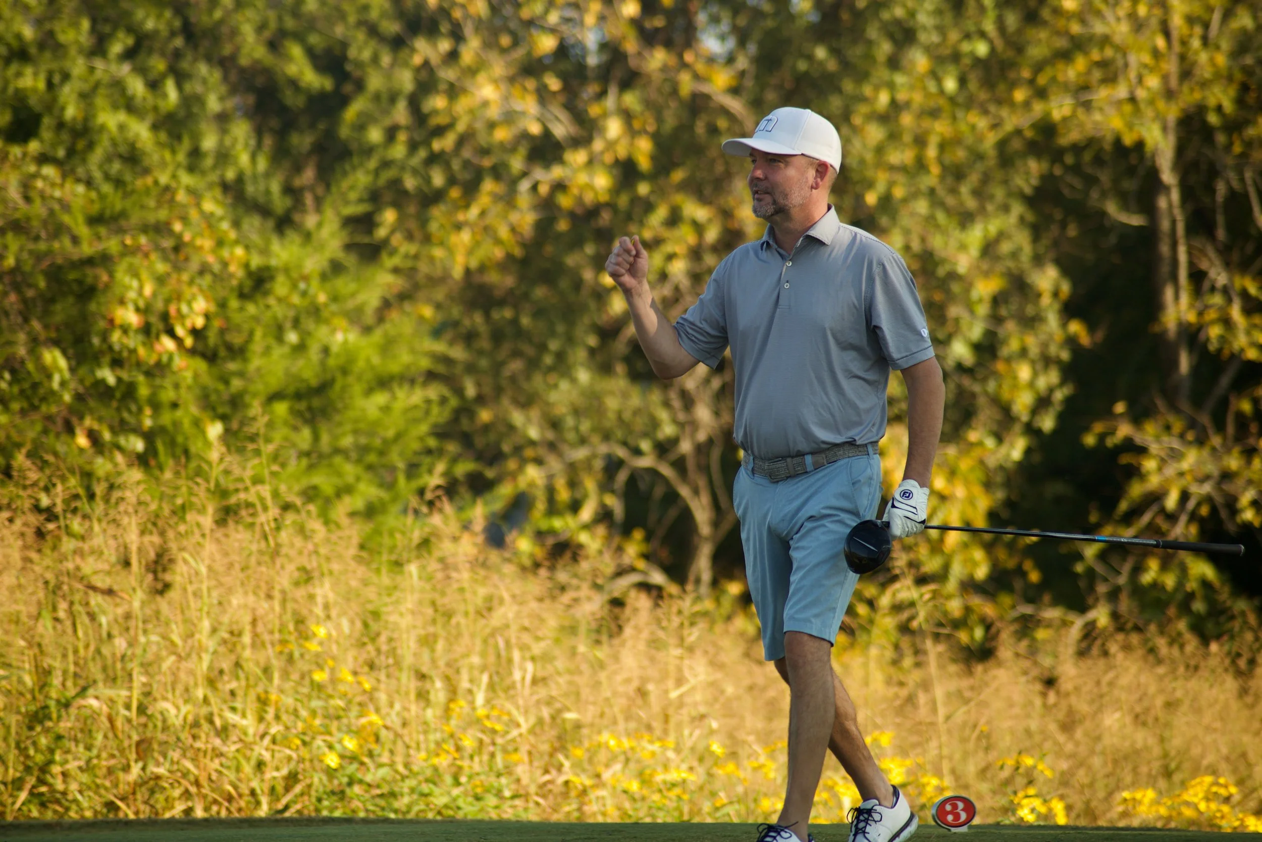 A man playing golf on a course surrounded by trees with yellow and green foliage. He is wearing a white cap, a light gray polo shirt, and blue shorts, holding a golf club and walking near a ball marked with the number 3.