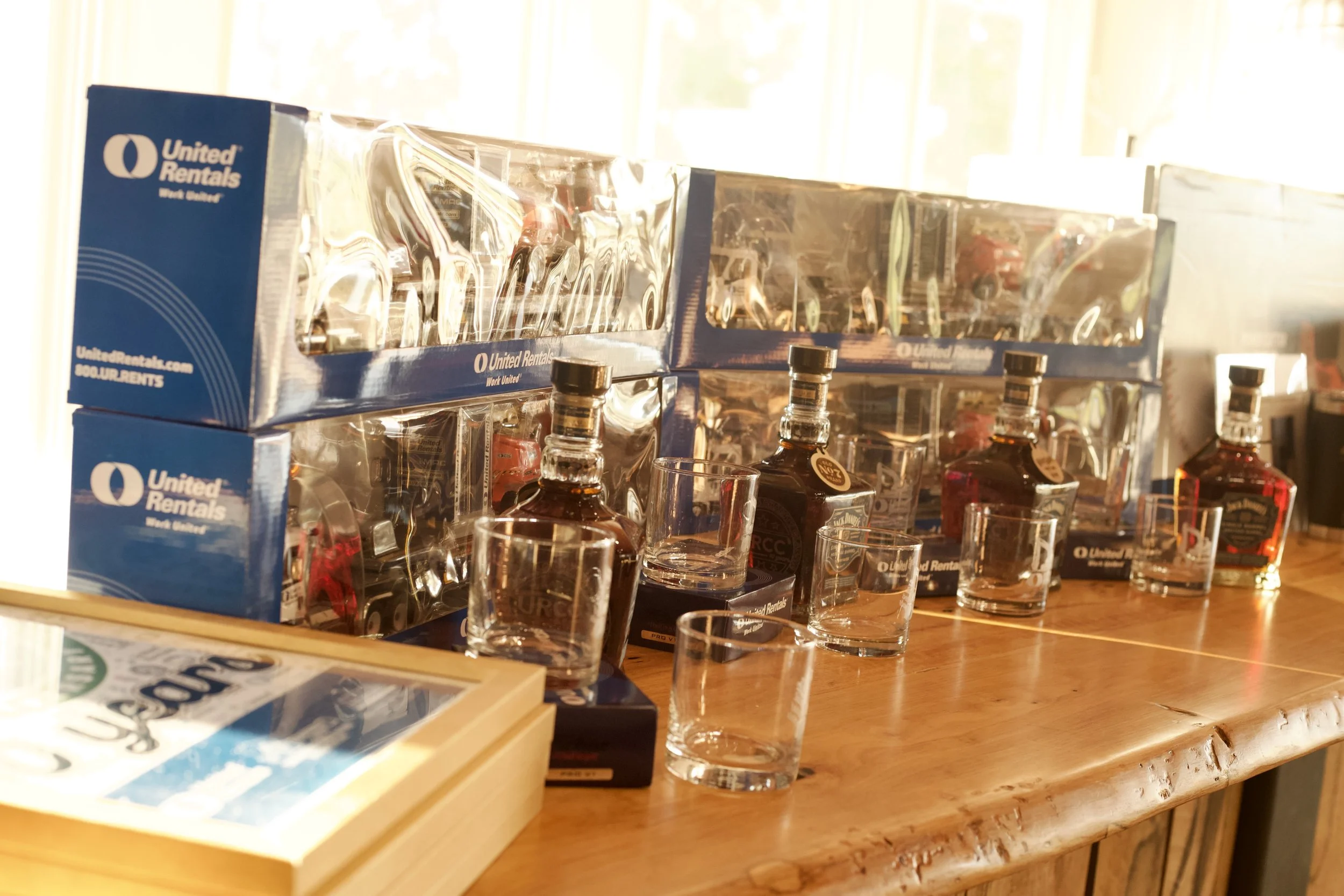 Boxes of toy cars and bottles of liquor on a wooden table with glasses in the foreground.