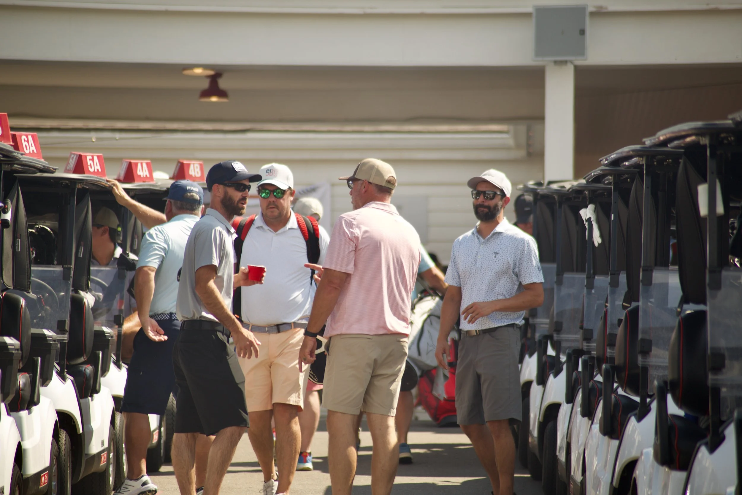 Group of men standing and conversing on a golf course or golf club parking lot, with golf carts parked on either side.