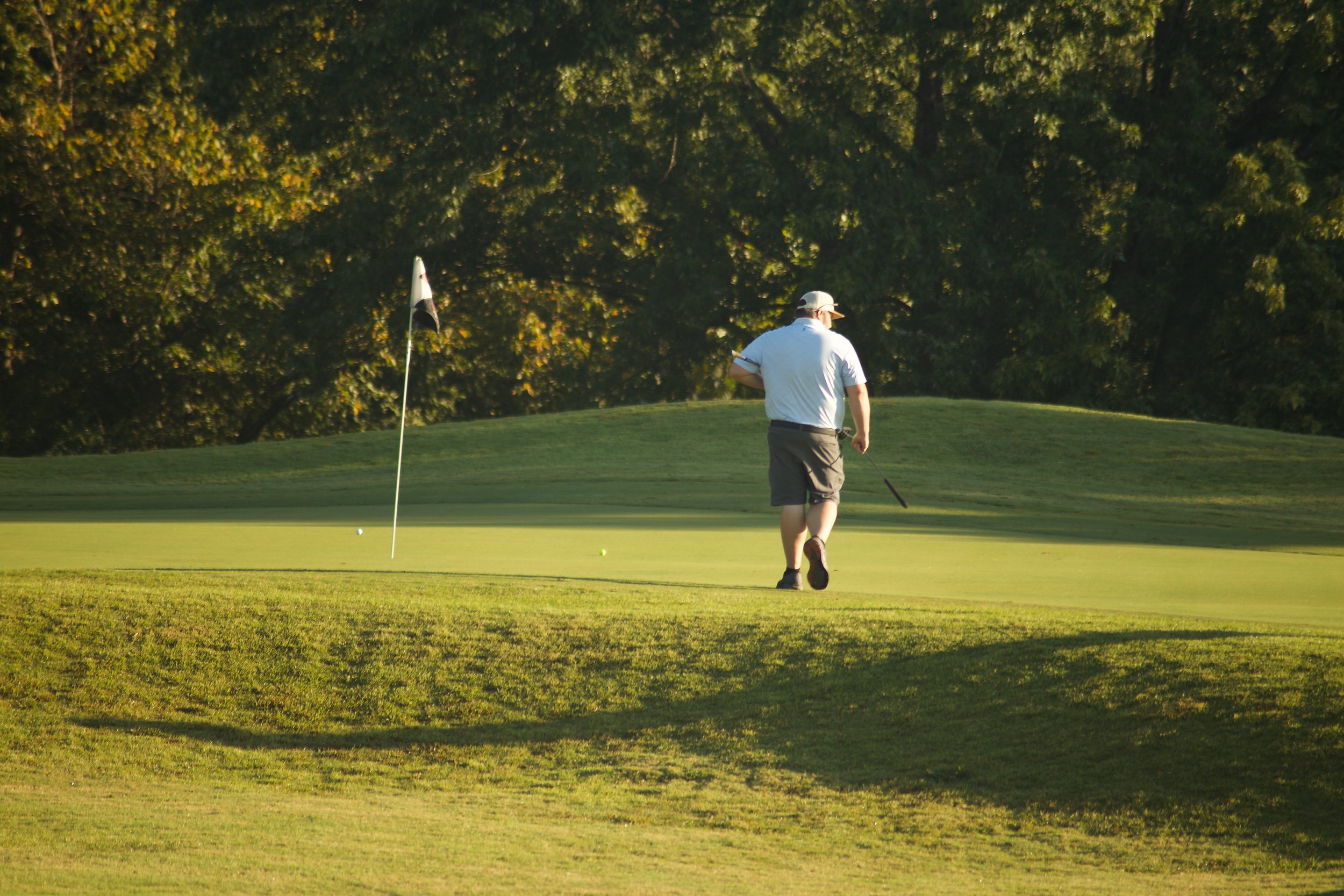 A man playing golf on a lush green golf course during late afternoon, preparing to putt a golf ball near the hole marked by a flagstick.