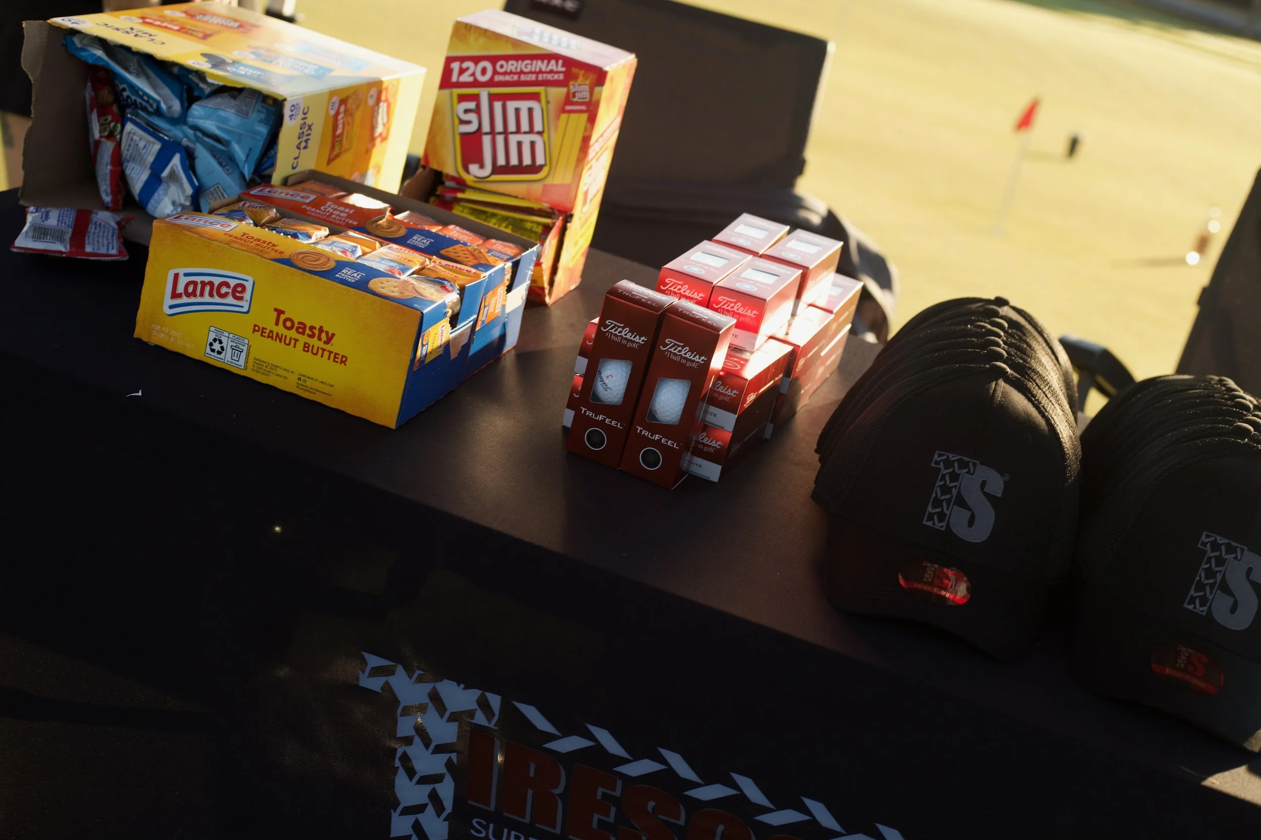 Open box of Lance snack packs, boxes of Slim Jim snacks, and boxes of Titleist golf balls on a black table outdoors with sports equipment nearby.