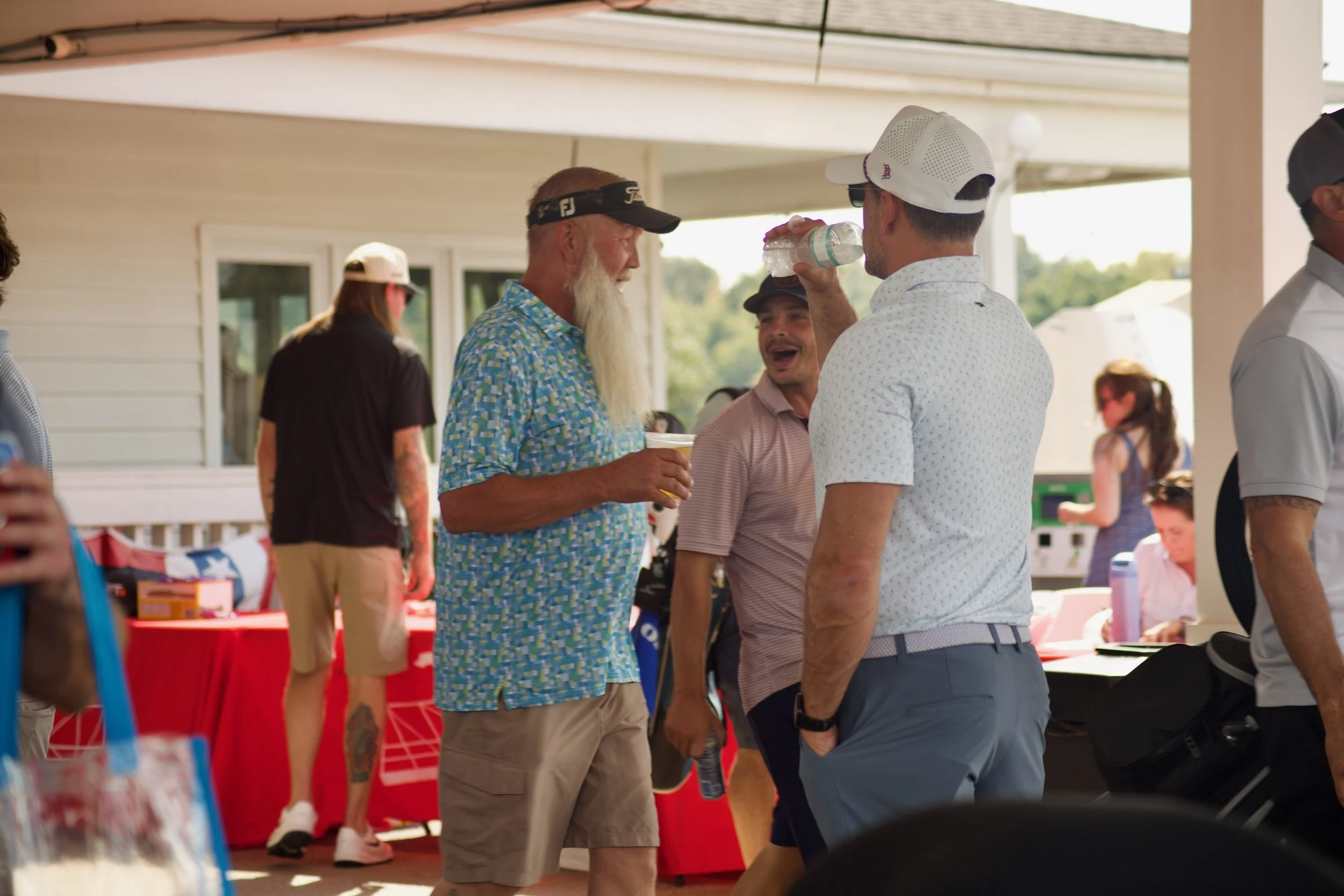 Group of men talking at an outdoor event, drinking water, standing near a red table with items for sale, in a sunny setting with a house in the background.