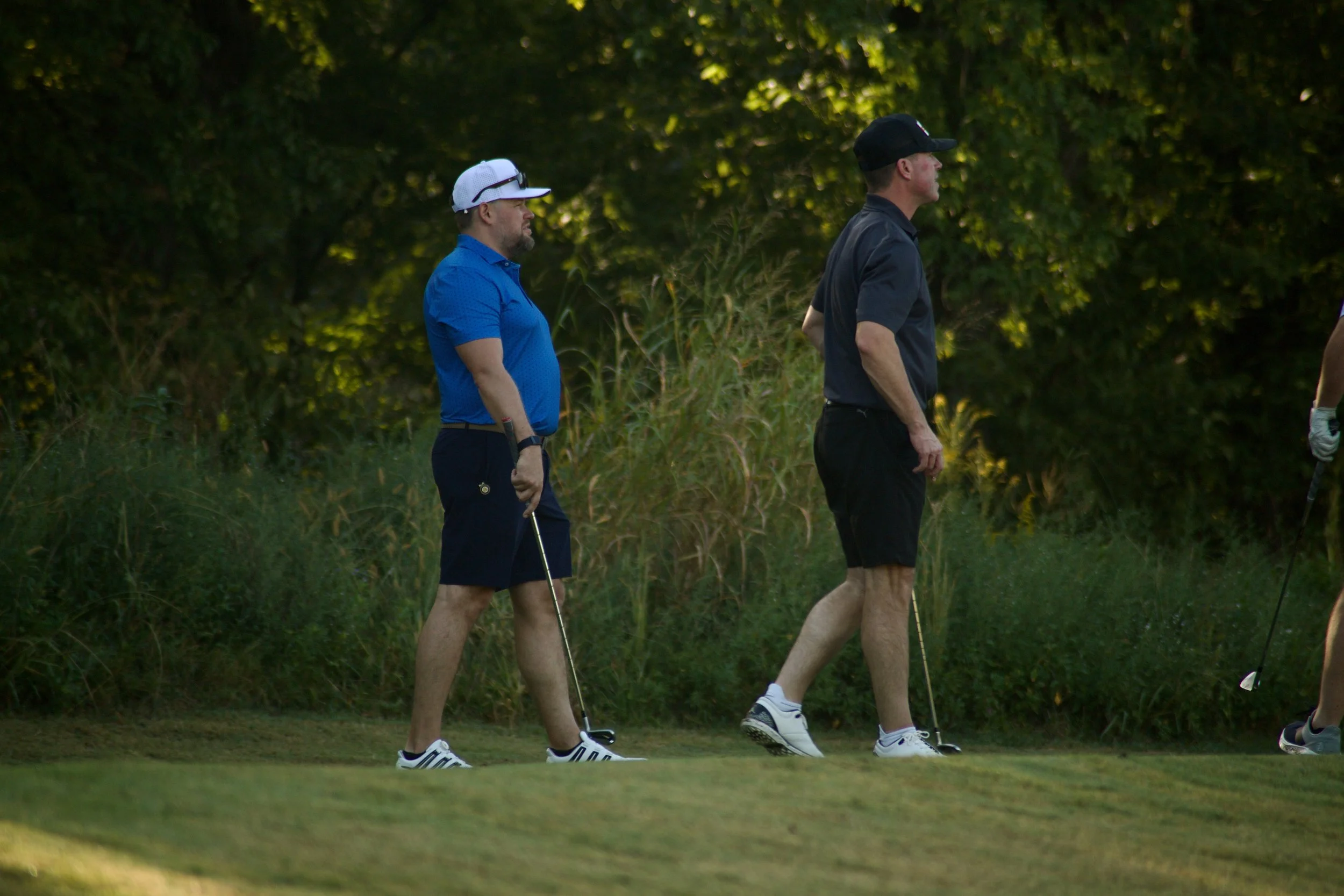 Two men walking on a golf course during daylight, one dressed in a blue shirt and white cap, the other in a black cap and dark shirt, with greenery in the background.