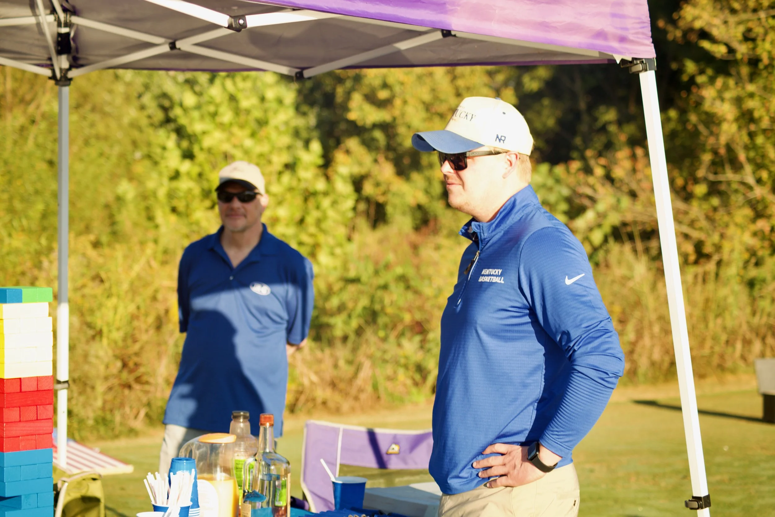 A man in sunglasses and a Kentucky basketball cap stands outdoors at a booth with drinks and supplies under a purple canopy, with two other men in blue shirts nearby.