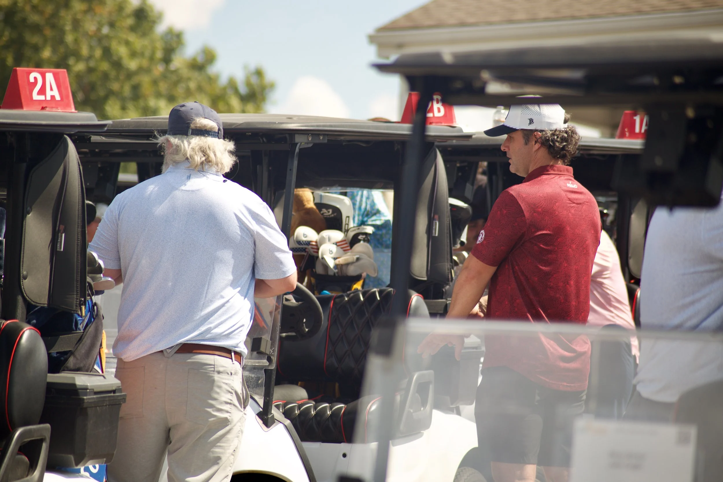 Two people, a man and a woman, standing beside golf carts filled with golf clubs at a golf course on a sunny day, with trees and a building in the background.