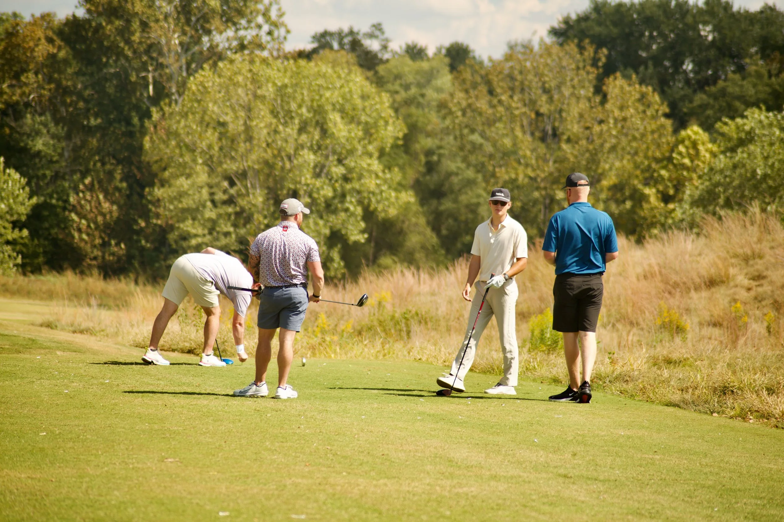 A group of five people playing golf on a sunny day in a grassy area with trees in the background.