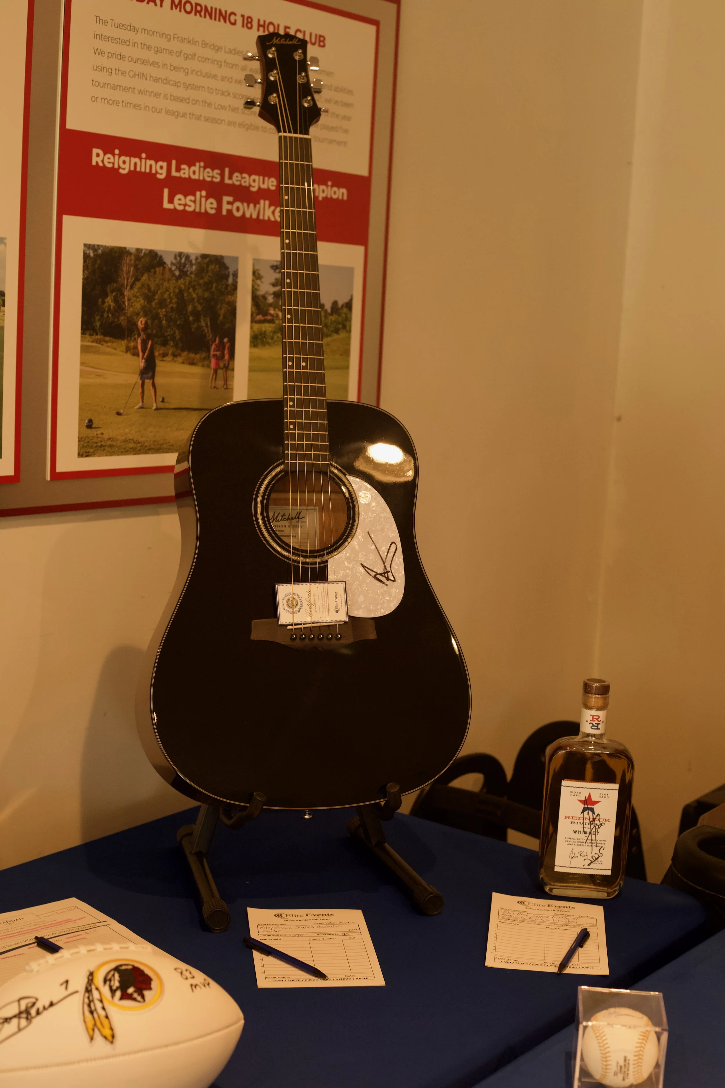Display of sports memorabilia including an autographed black acoustic guitar, signed football, baseball, and various papers and awards on a table, with a poster in the background mentioning a golf league champion named Leslie Fowke.