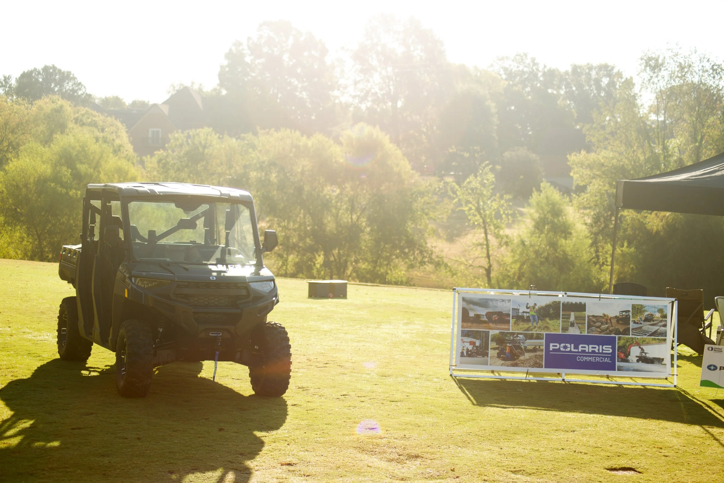 A Polaris commercial utility vehicle parked on a grassy field at an outdoor event, with a display board featuring images and the Polaris logo in front of the vehicle.