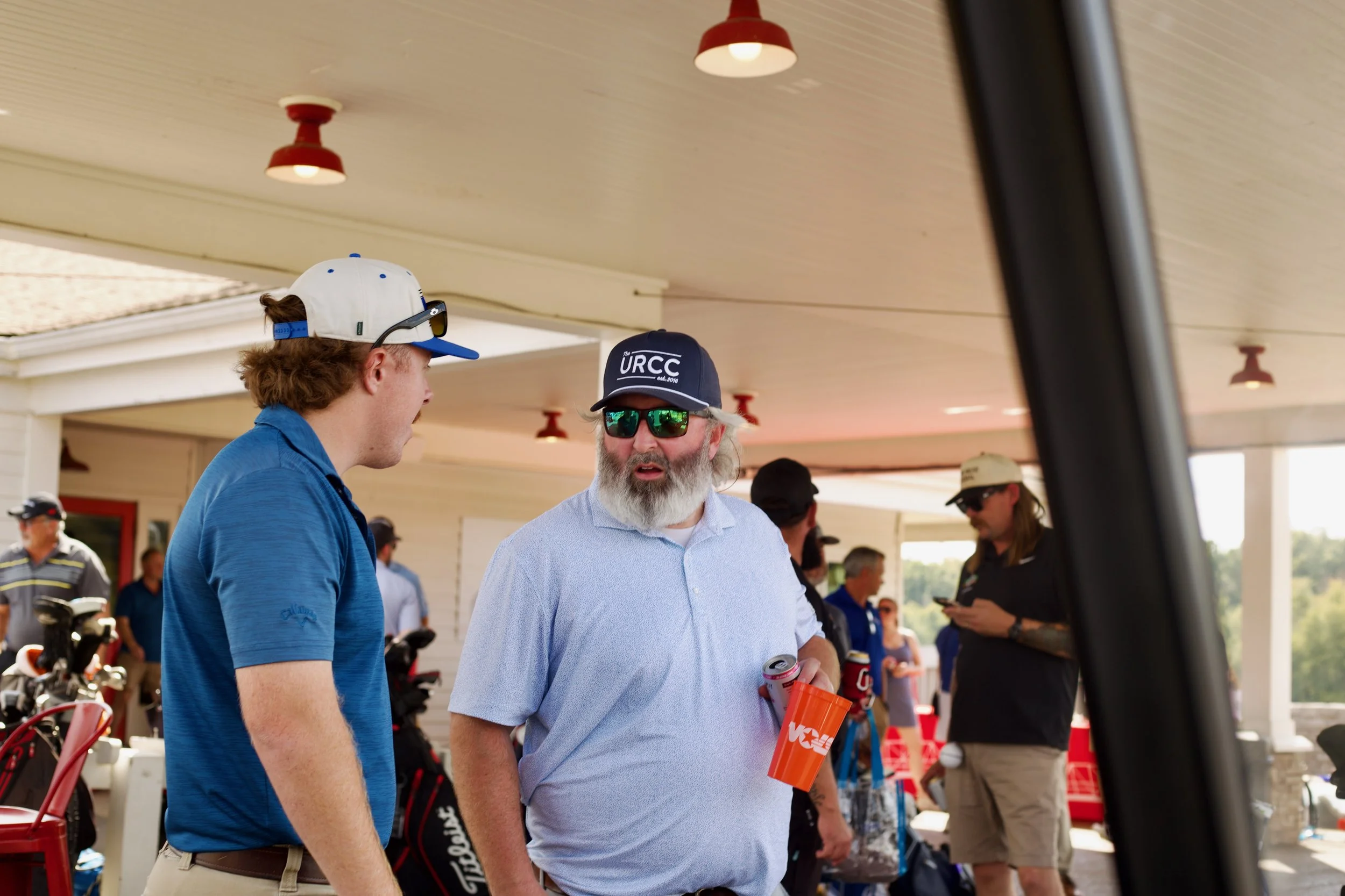 Two men engaged in conversation at an outdoor gathering, with golf clubs and equipment visible. One man has a beard, sunglasses, and a cap, while the other man has brown hair, and a cap with sunglasses resting on it.