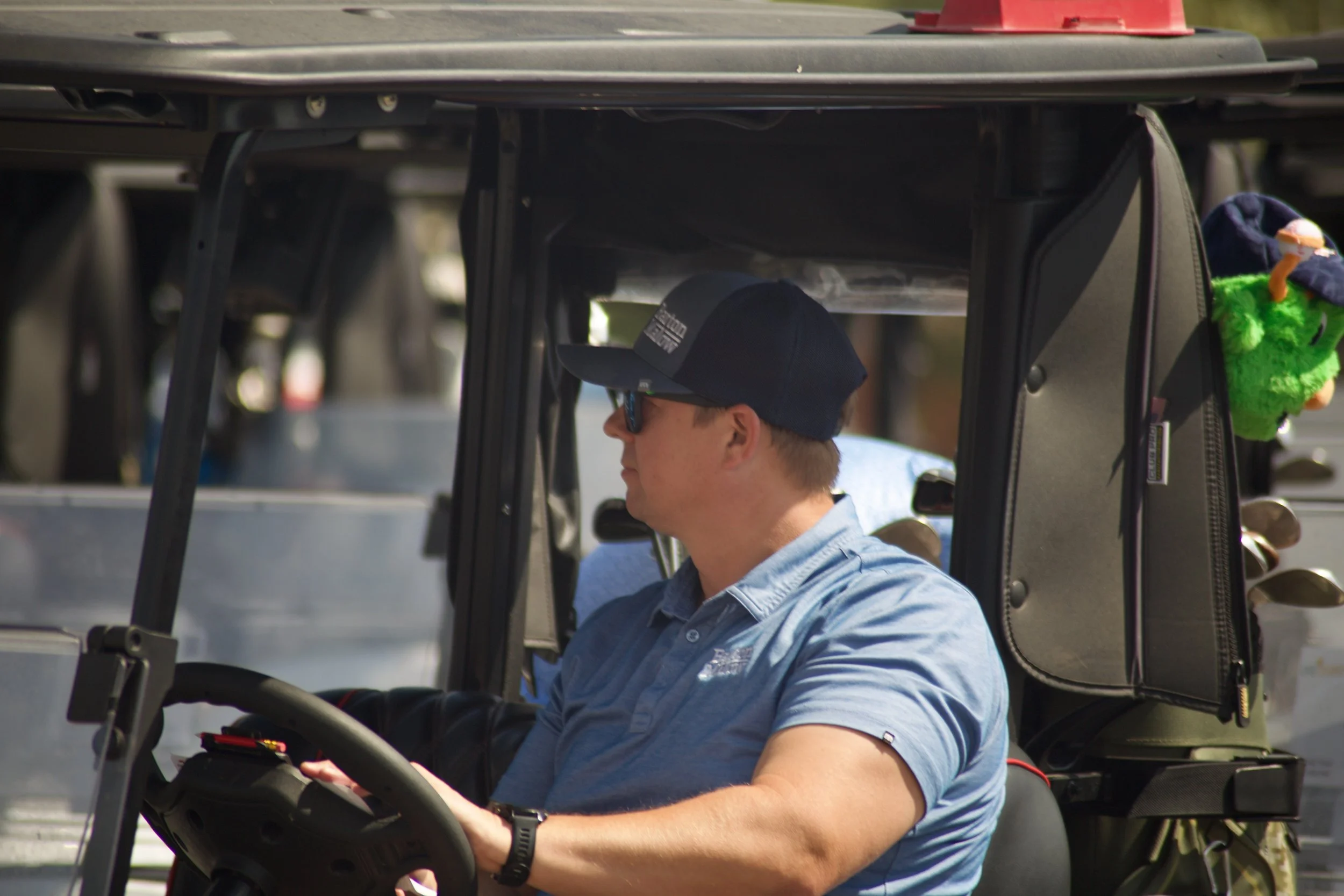 A person with light skin, wearing sunglasses, a baseball cap, and a light blue shirt, is sitting in a golf cart, looking at a device in their hands. The golf cart has a black roof and a small hanging plush toy on the side.