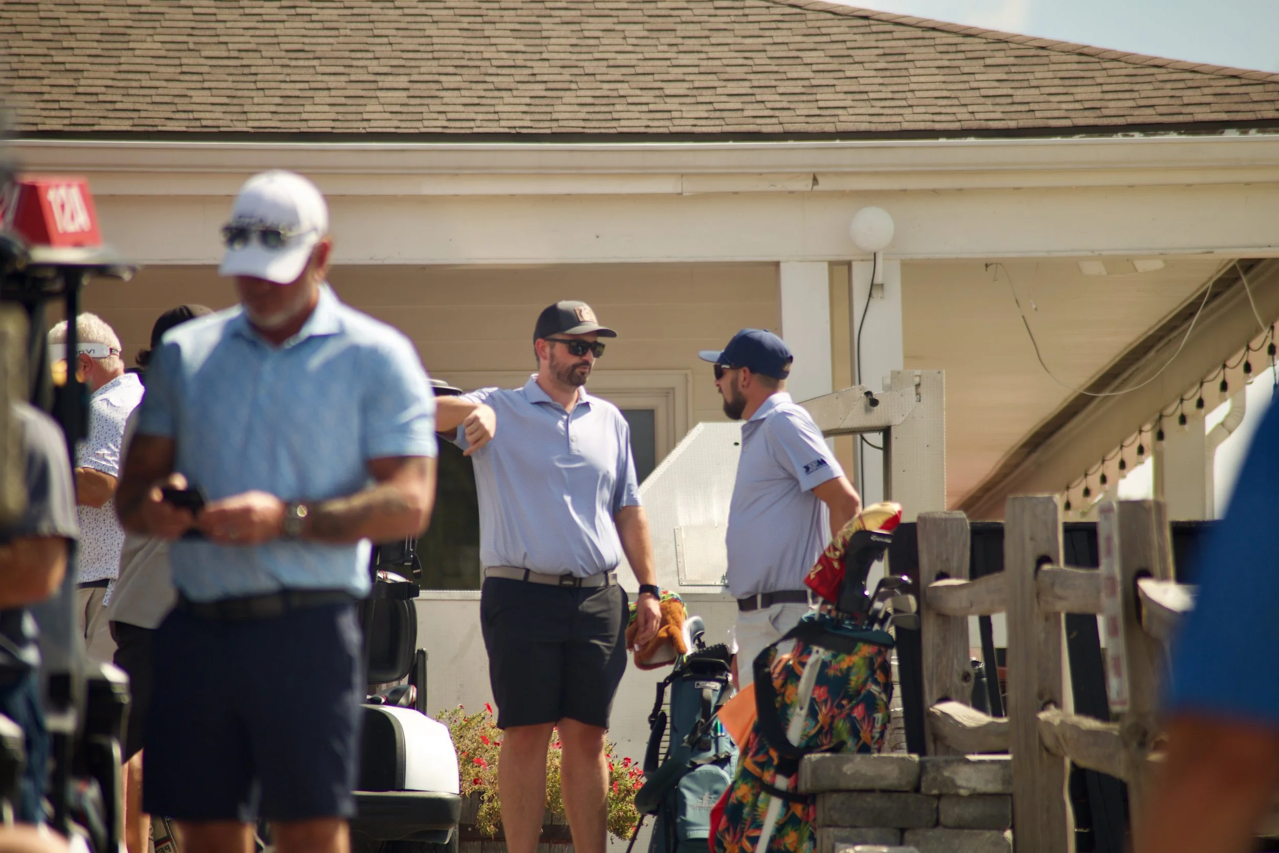 People gathered outdoors near a house with a brown shingle roof, dressed in casual summer clothing, some wearing sunglasses and hats, with golf bags and equipment visible.