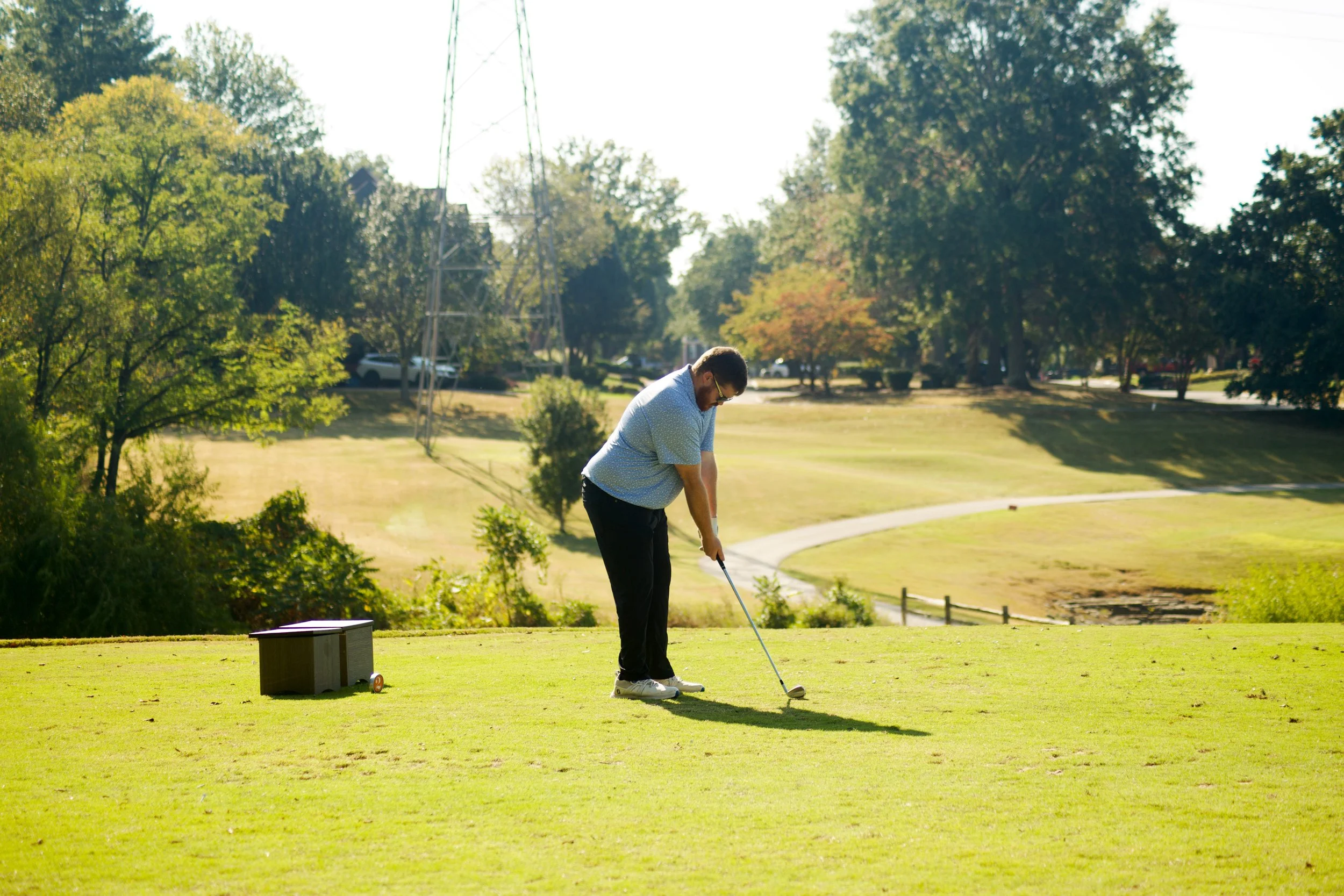 A man playing golf on a lush green course during daytime, with trees and power lines in the background.