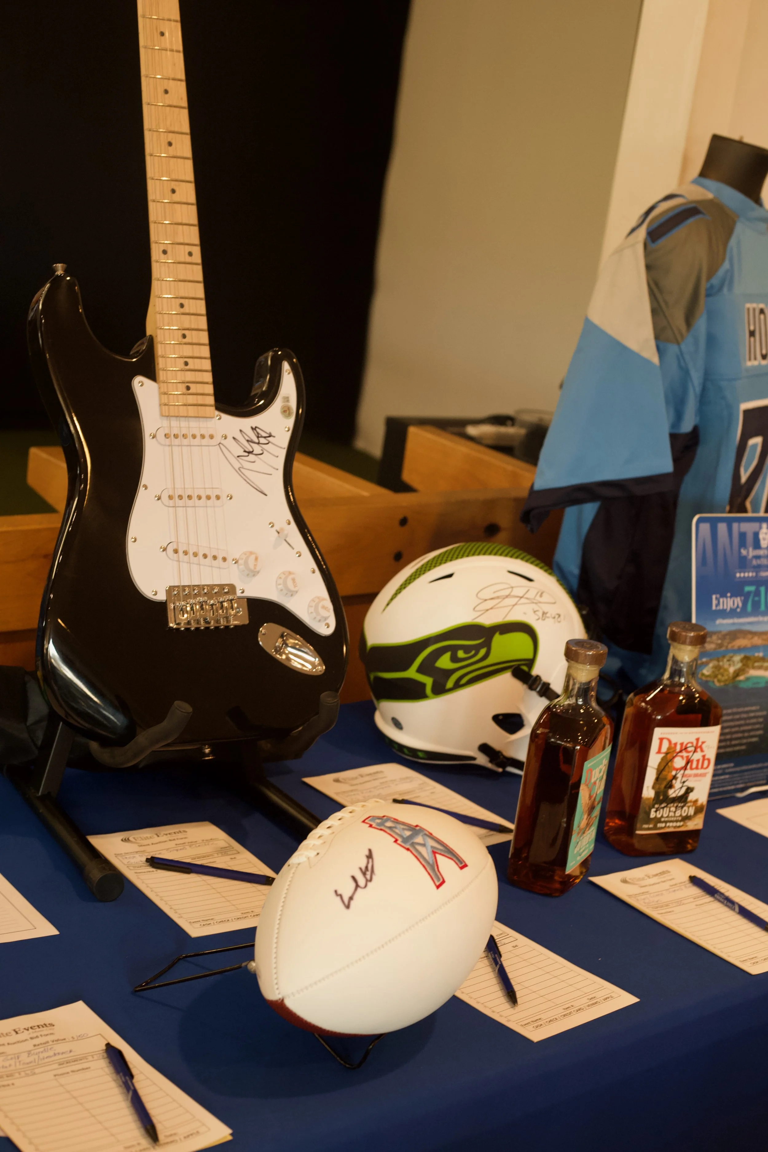 Display table with signed footballs, a motorcycle helmet with a stickers of a black and yellow face, a motorcycle helmet with a red and black logo, bottles of alcohol, and some papers and pens.