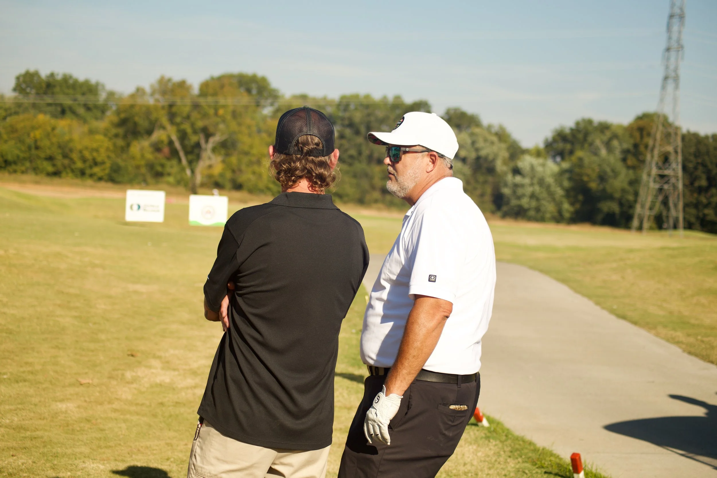 Two men stand on a golf course talking, with backup signs and trees in the background.