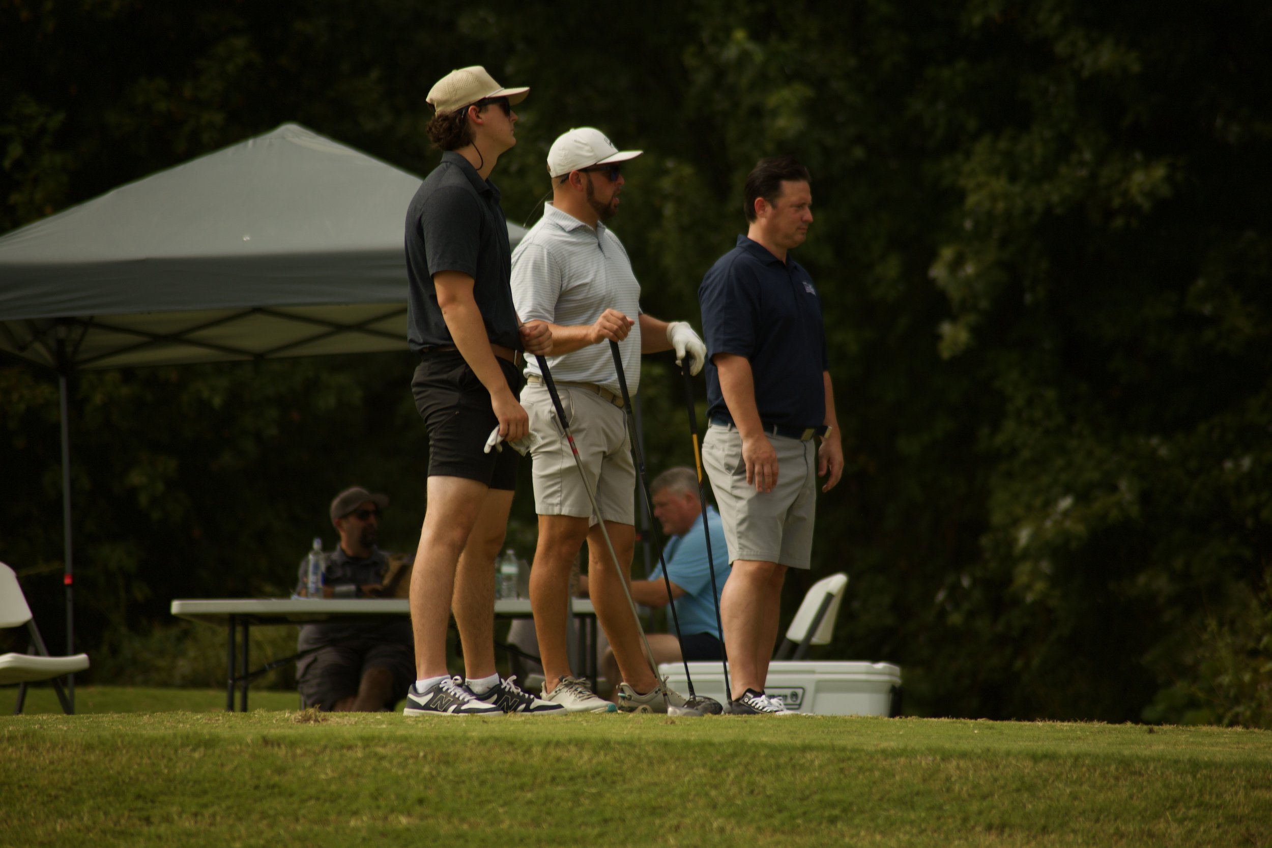 Three men standing on a golf course, preparing to putt, with two sitting at a table in the background.