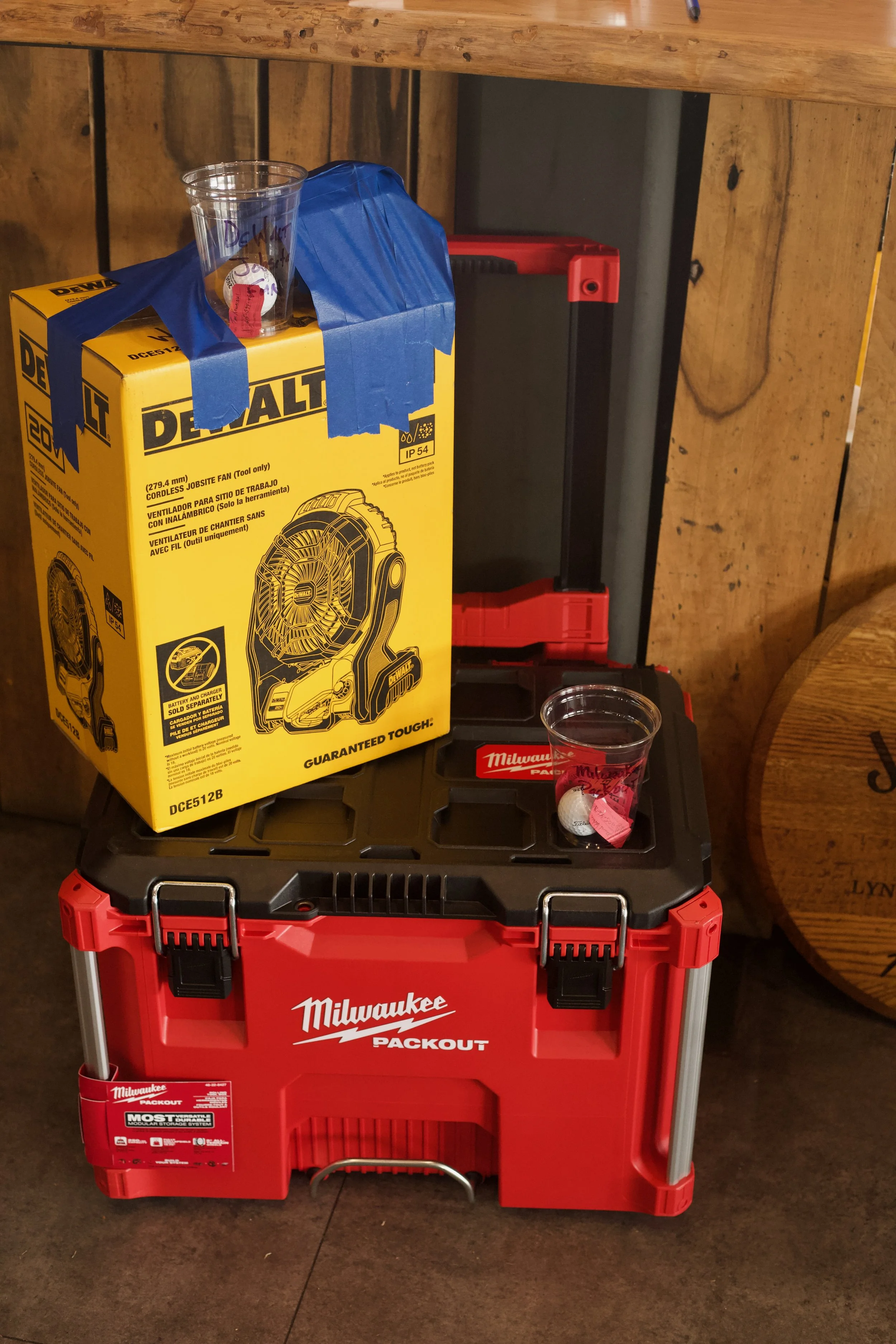 A red Milwaukee Packout toolbox stacked with a black base, with a yellow Dewalt box and a clear plastic cup placed on top, against a wooden background.
