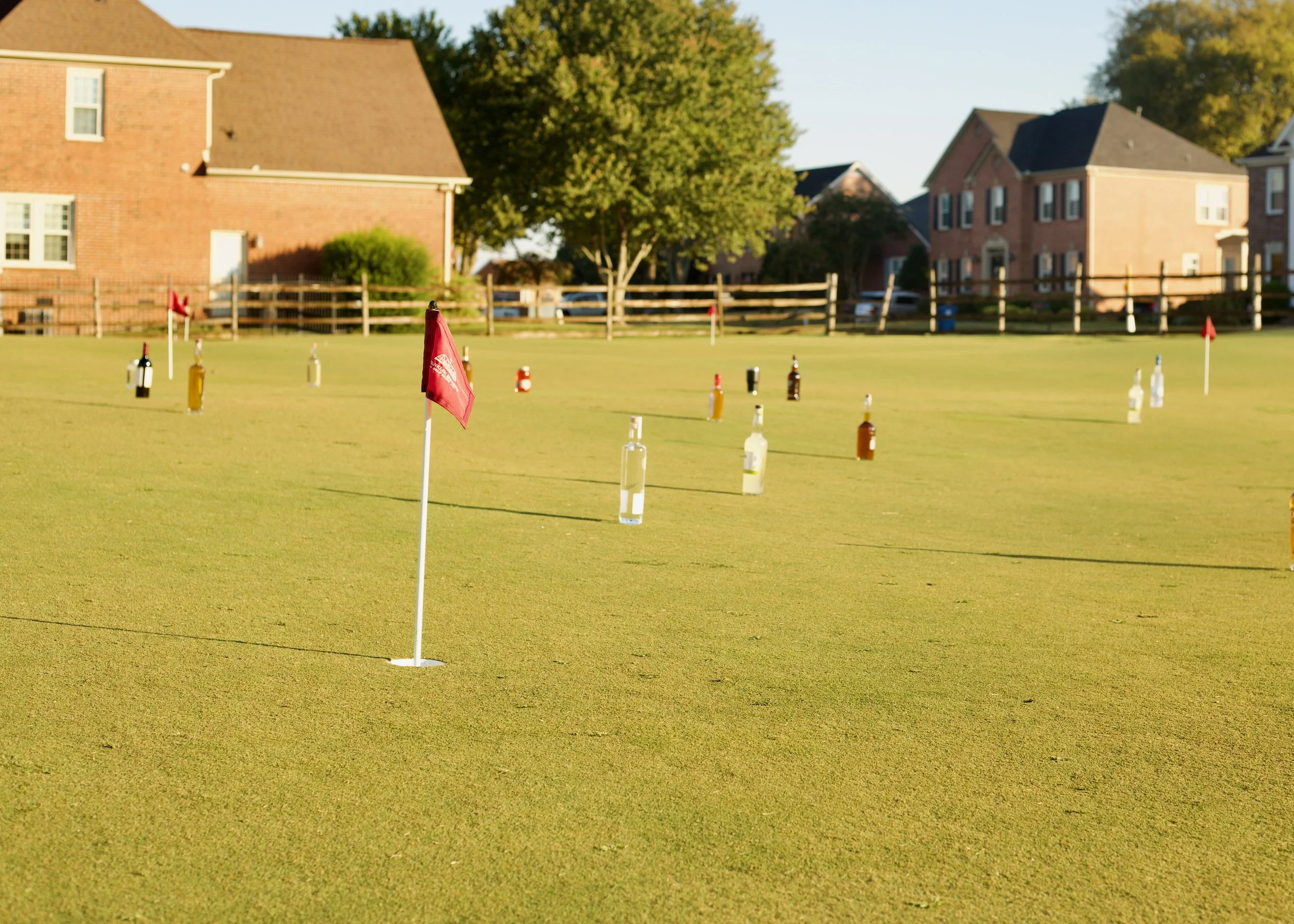A putt golf green with flags and bottles, residential houses, green trees, and clear sky in the background.