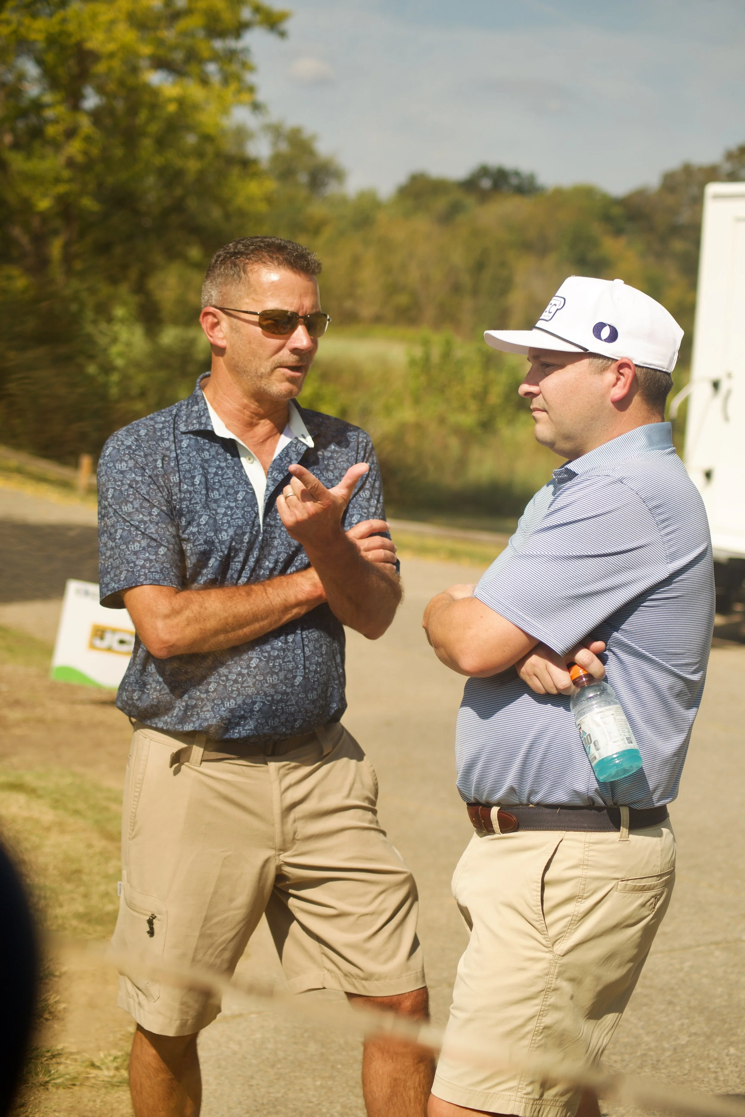 Two men stand outdoors in a sunny, rural setting, engaged in conversation. One man points while speaking, the other man listens with arms crossed, holding a water bottle. Both are wearing casual shirts and shorts; one wears sunglasses and a white cap