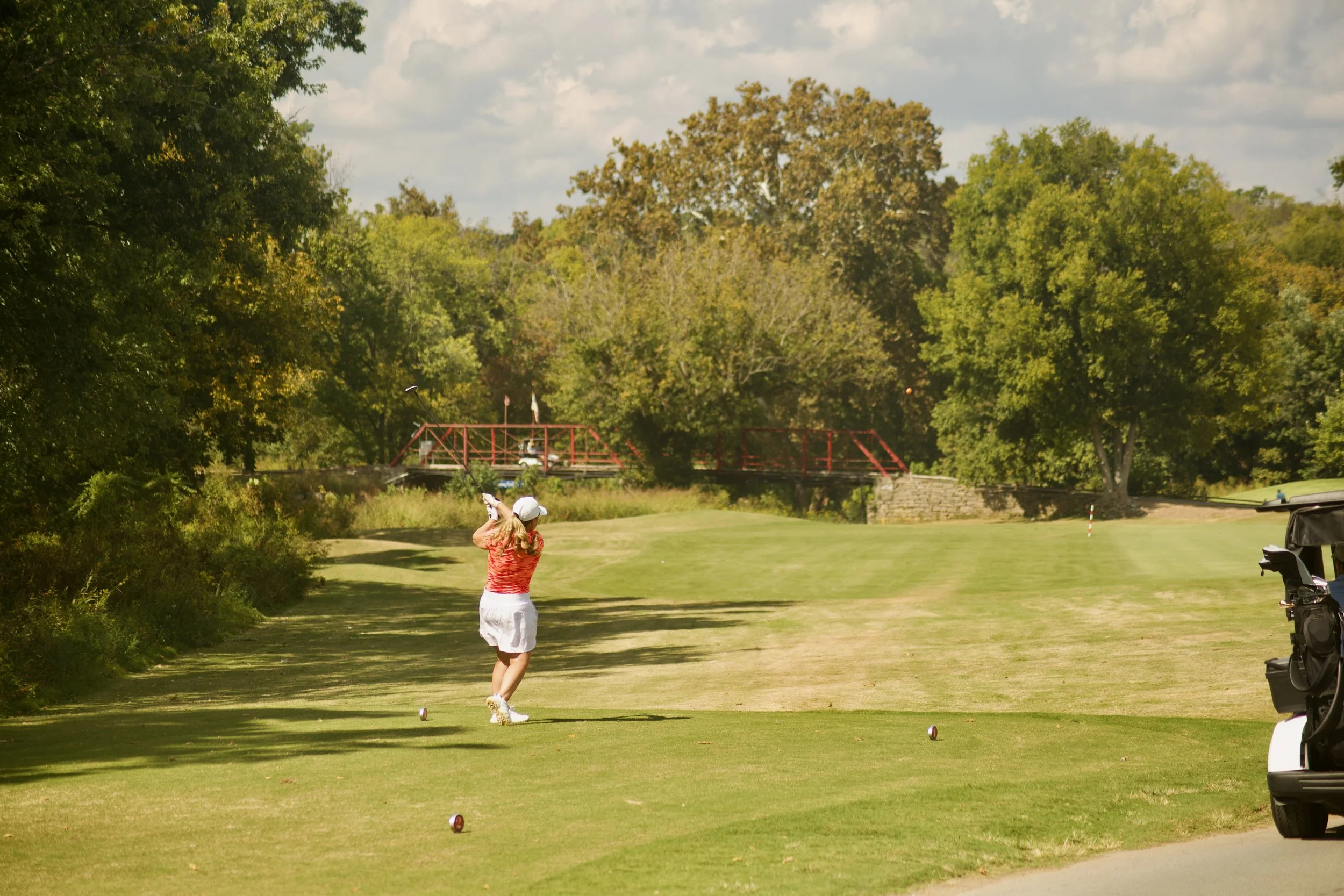A woman in a red patterned shirt and white skirt playing golf on a lush green golf course. She is preparing to hit a golf ball, with a golf cart nearby. The background features tall trees and a partly cloudy sky.
