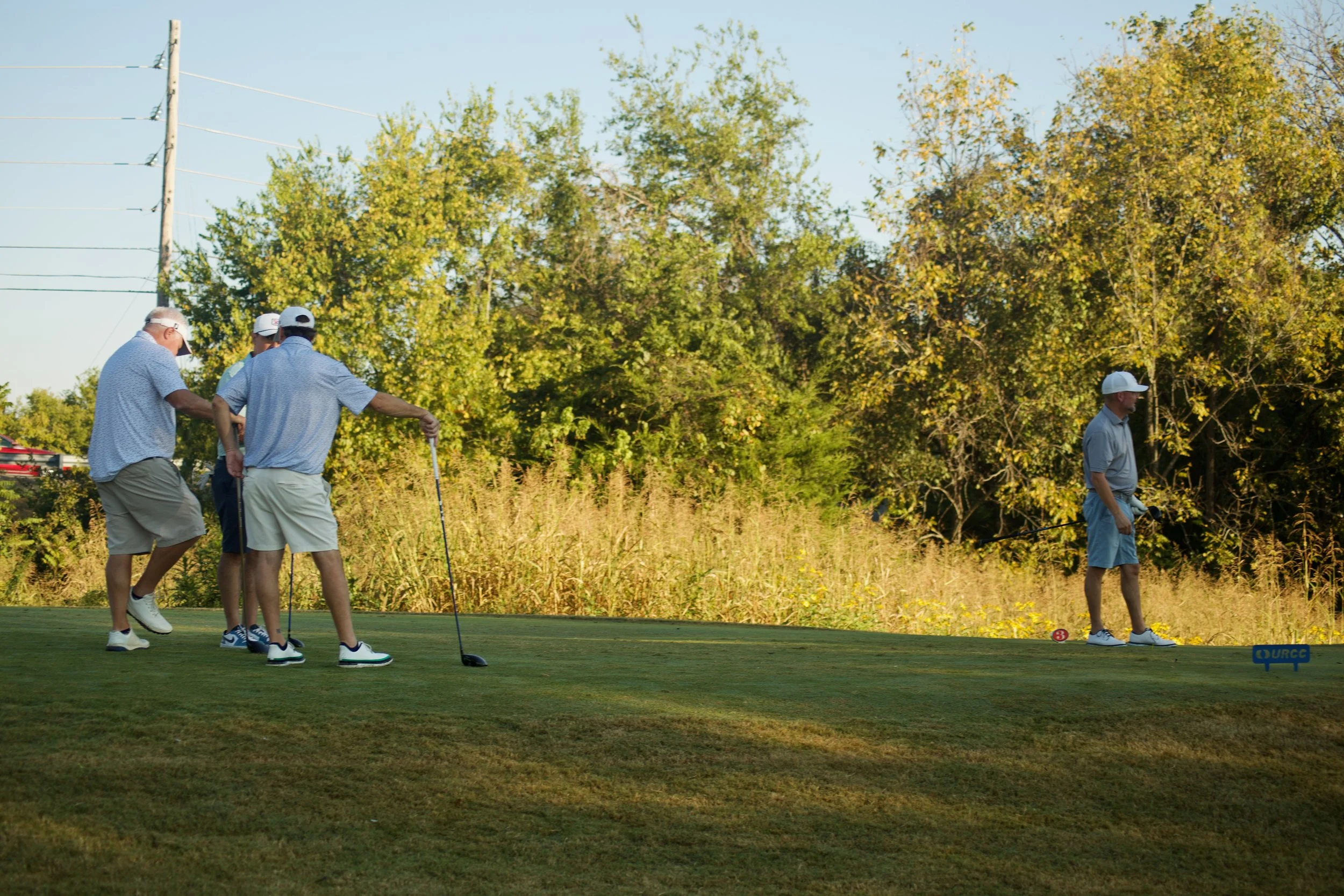 Four men on a golf course, one man preparing to hit a golf ball while others watch, surrounded by trees with autumn foliage.