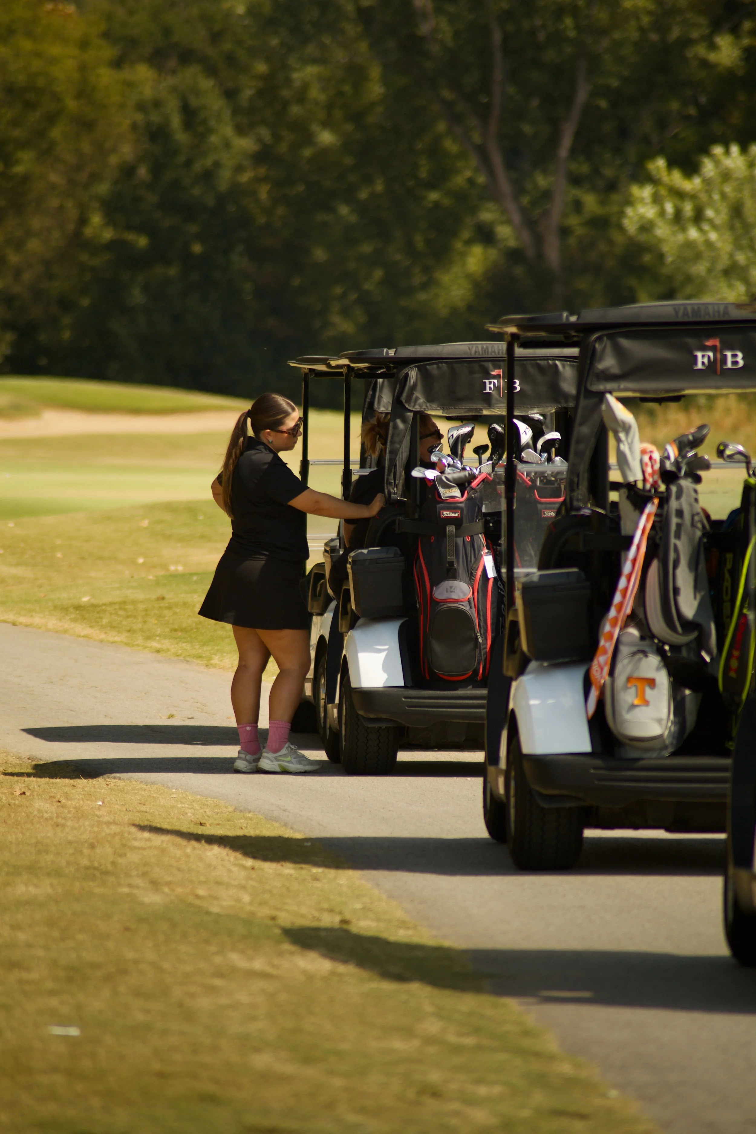A woman in a black dress and pink socks is adjusting a golf bag on a golf cart. The golf cart is parked on a golf course with trees in the background.