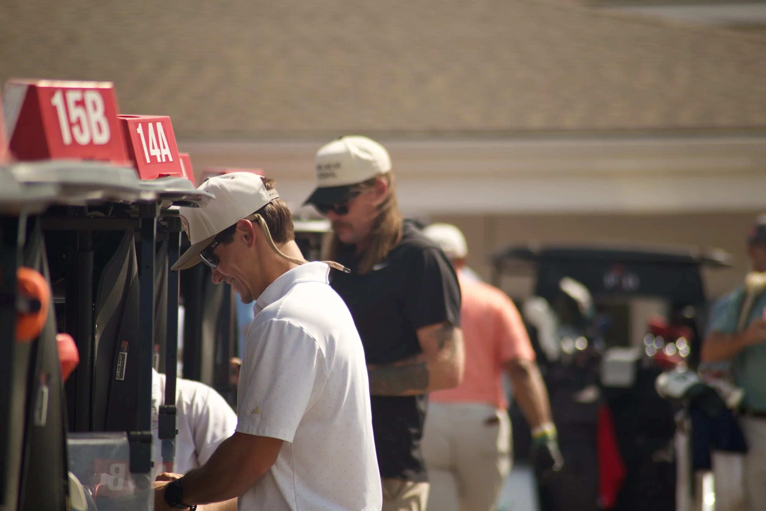 A row of golf bags with numbered signs, some people wearing hats and casual clothing visible in the background at an outdoor golf course or practice area.