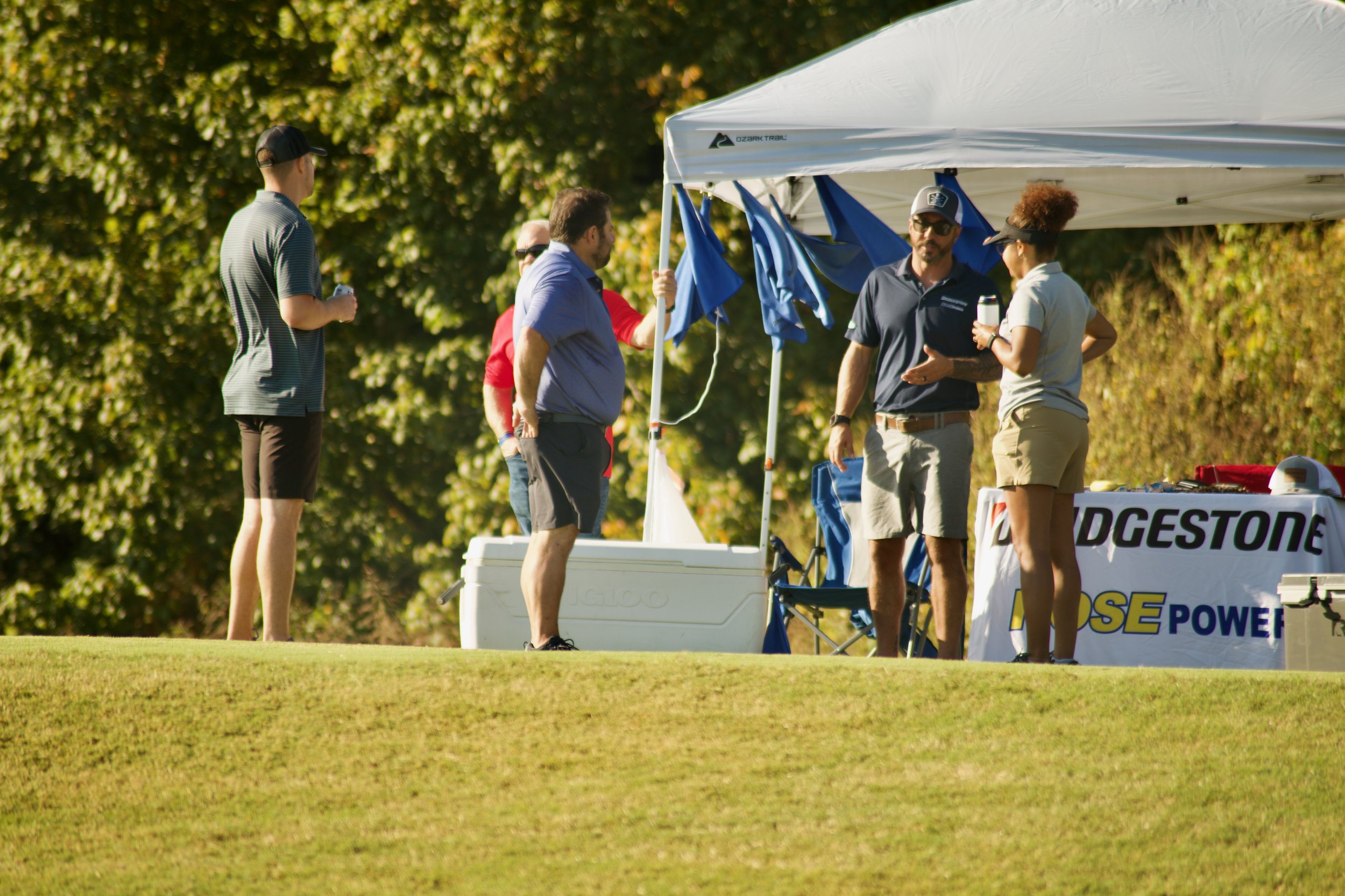 Group of people gathered under a white canopy at an outdoor event on a sunny day, with trees in the background.
