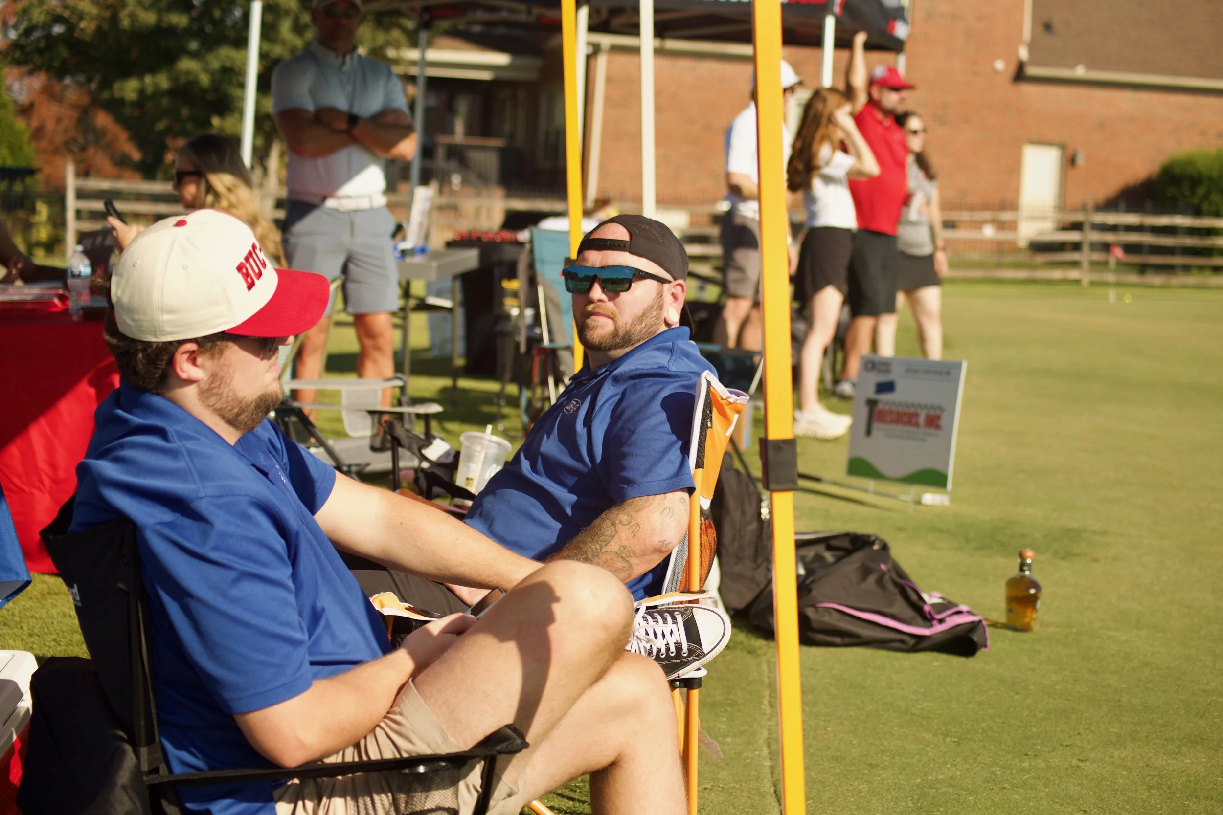 People sitting outdoors under a tent on a grassy field during an event, some engaged with their phones, others standing in the background.