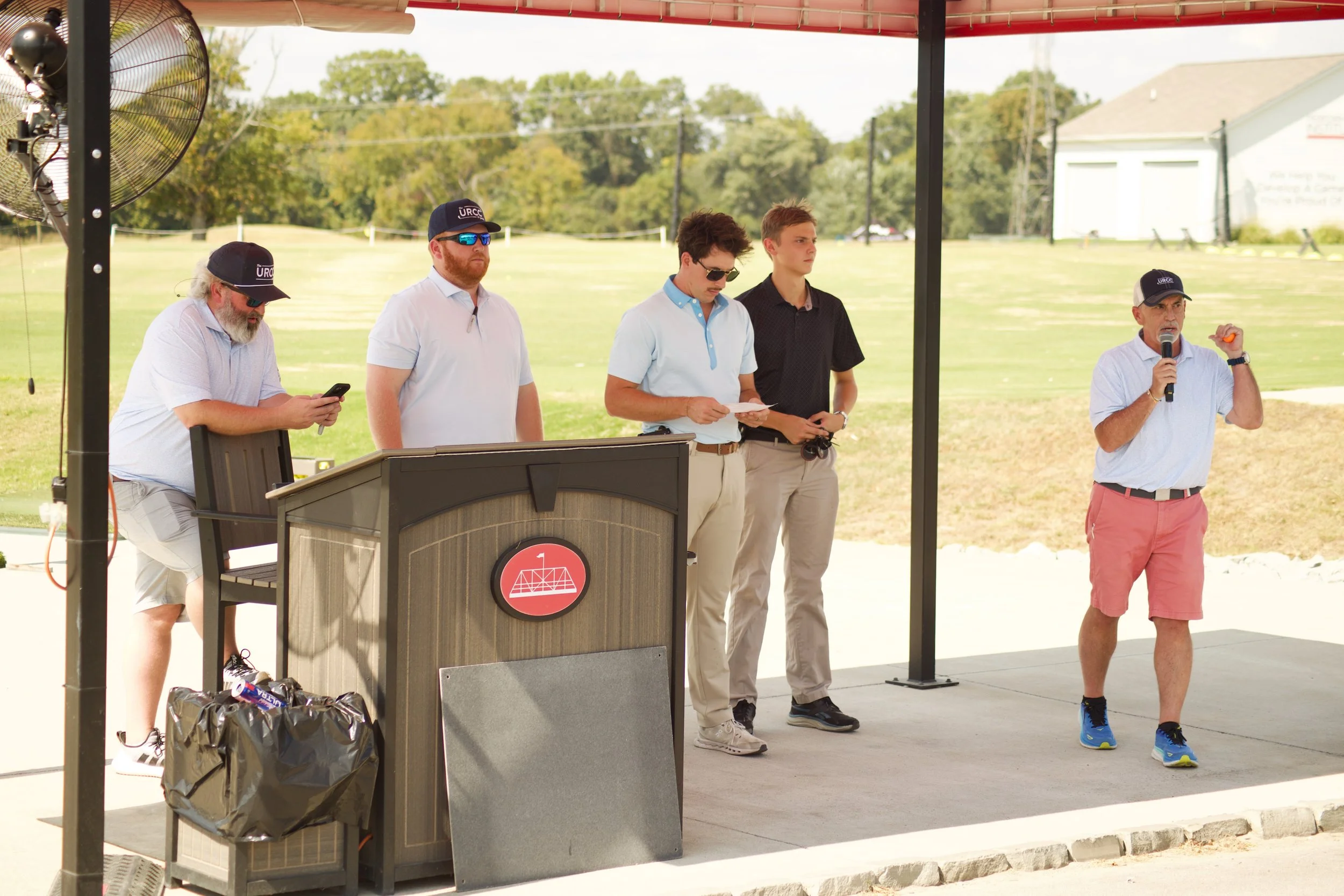 A group of five men standing outdoors, one man speaking into a microphone and others looking on, with a podium and two large fans nearby, at a sunny day at a park or sports field.