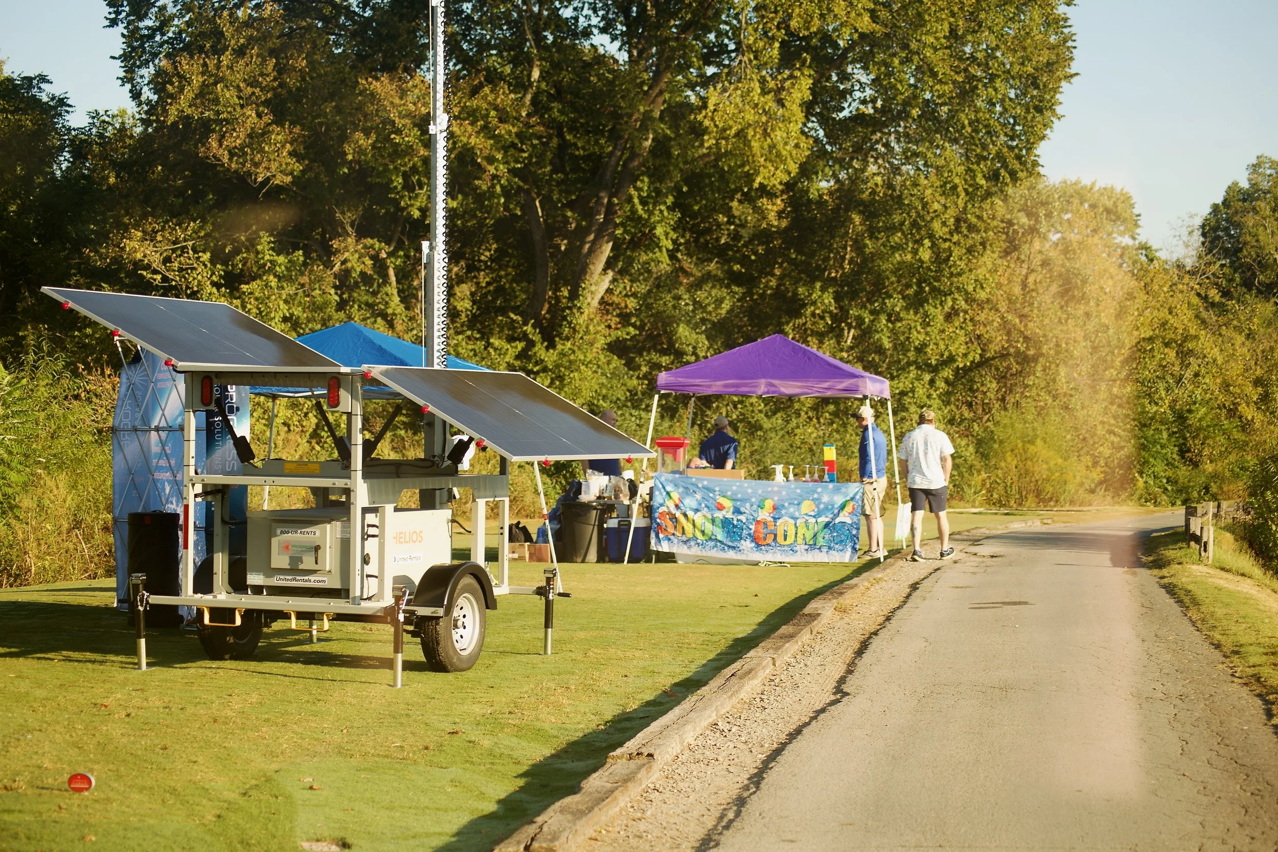 An outdoor scene with a trailer and a canopy tent at a small event near a wooded area, with people walking along a sidewalk.