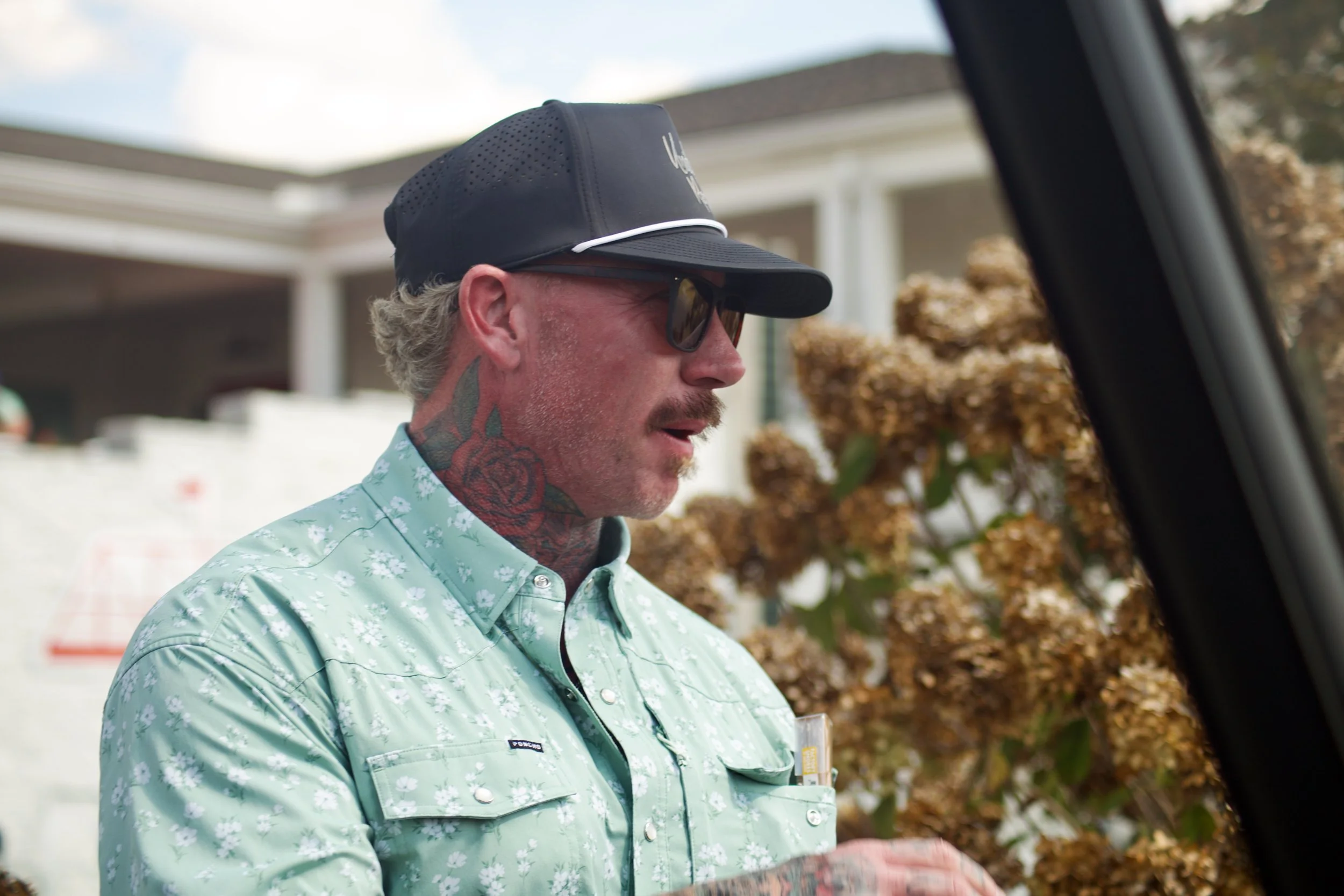 A man wearing sunglasses, a black cap, and a light green shirt with white floral patterns is standing outdoors. He has tattoos and is near a large pile of brown mulch.